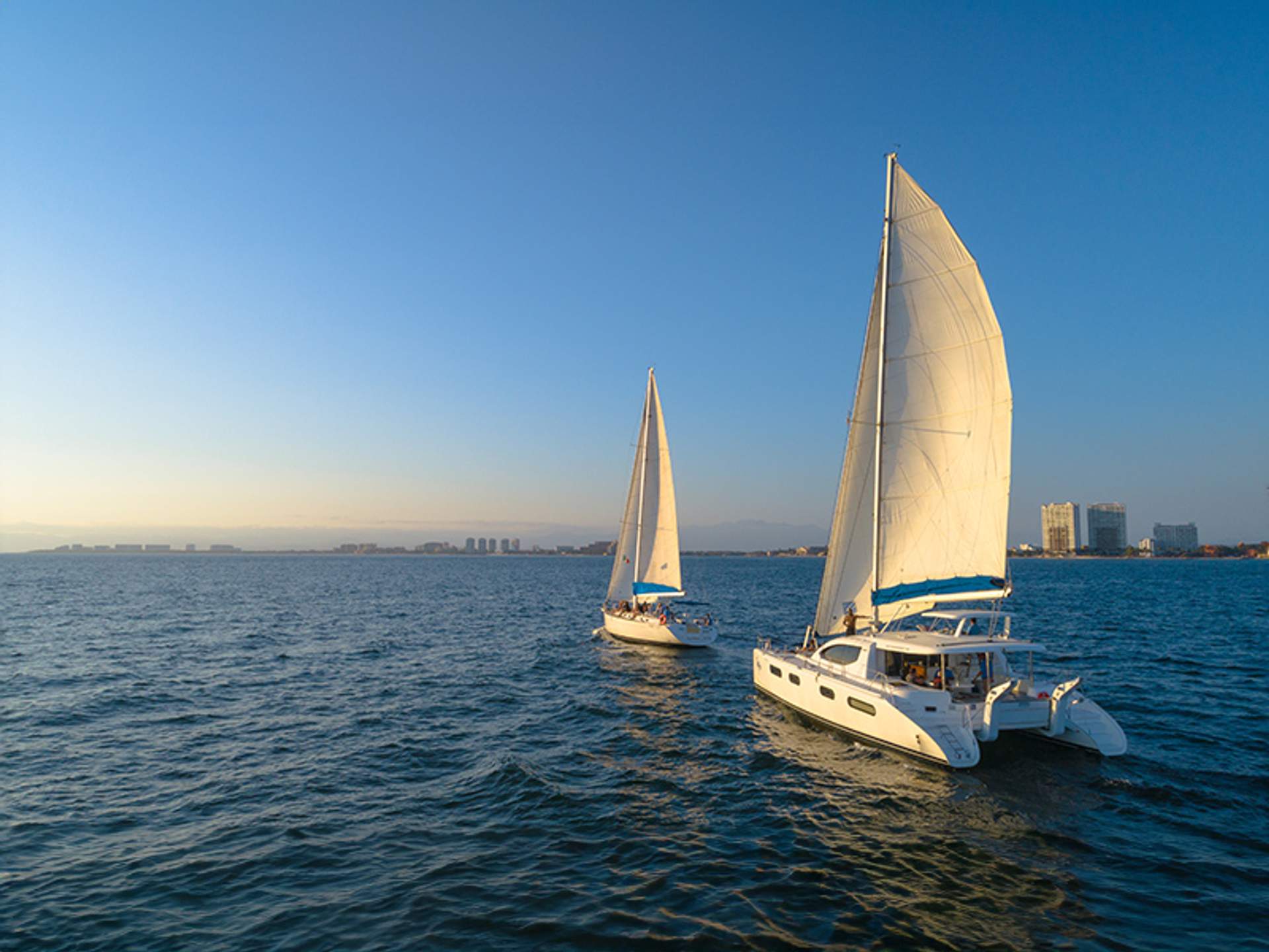 Dos veleros blancos desplegadas navegando cerca de la costa de Puerto Vallarta al atardecer, con edificios al fondo.
