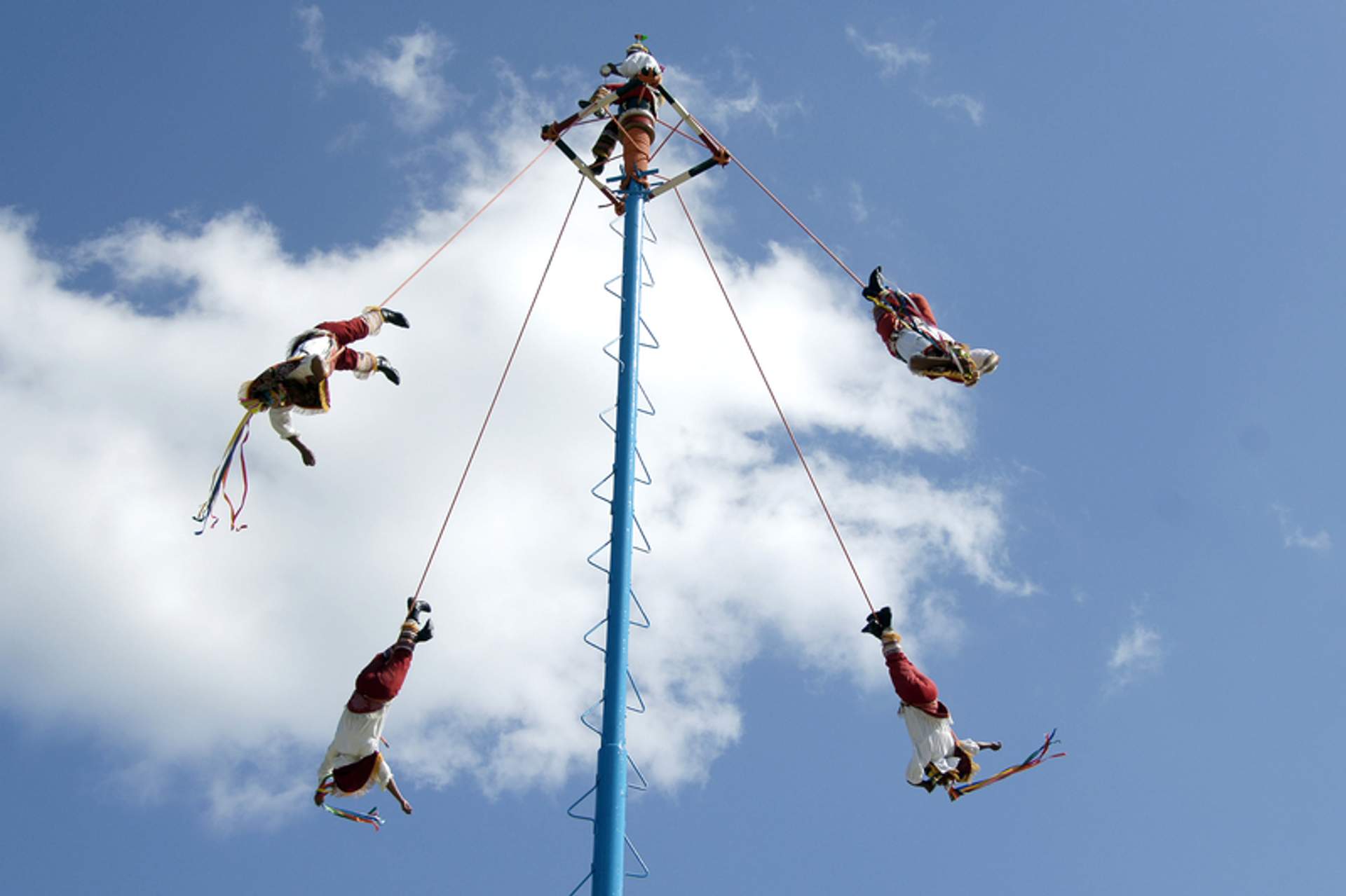 Performers known as "Voladores de Papantla" are suspended in mid-air, spinning around a tall pole against a blue sky with clouds.