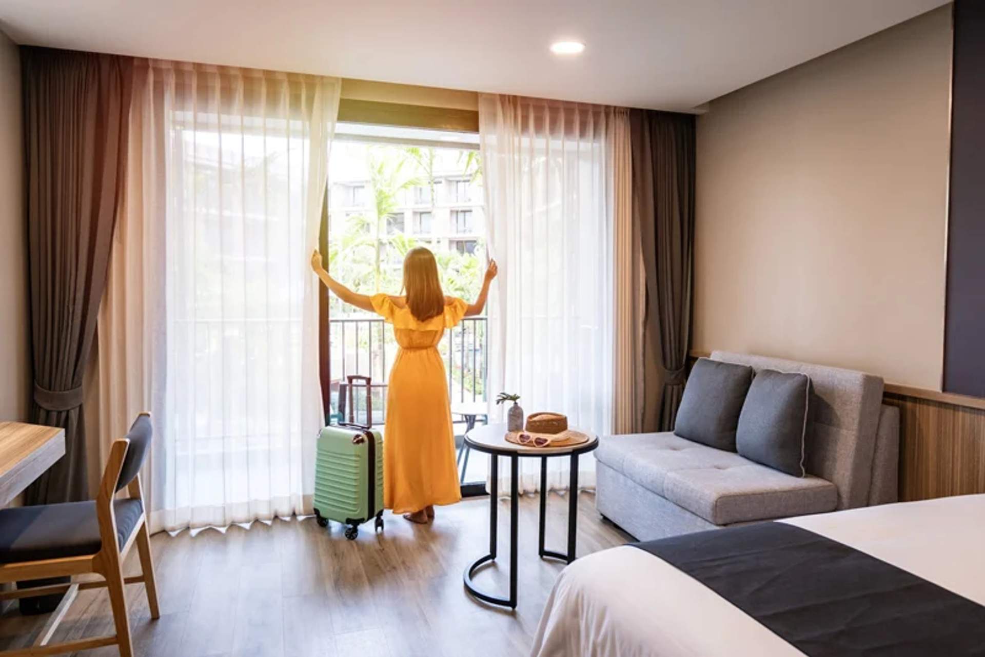 A woman in a yellow dress standing by a large window in a hotel room, opening the curtains. A green suitcase is beside her, and the room features a bed, a sofa, and a table with snacks.