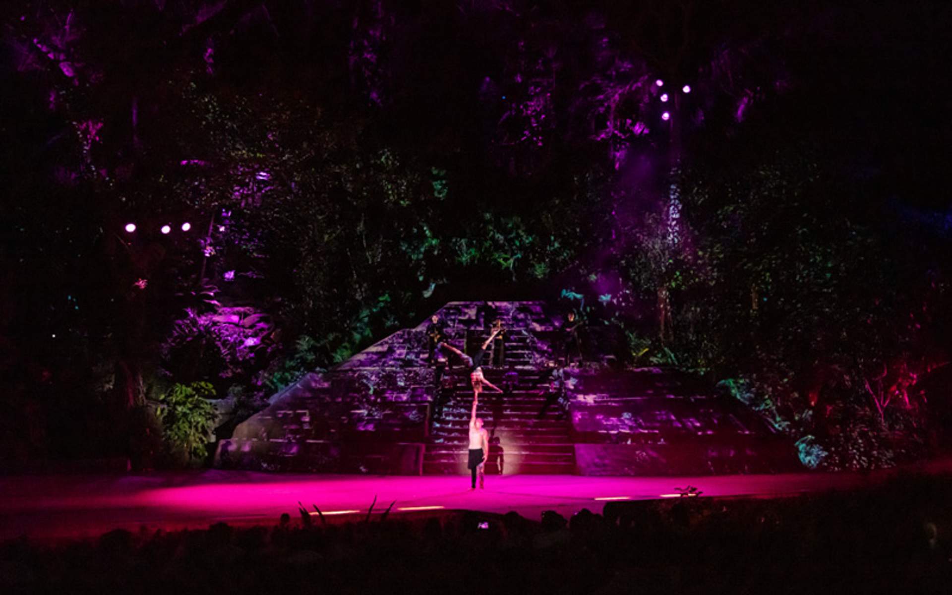Performers on a lit stage with ancient stone steps in the background, surrounded by vibrant lights and foliage at night.