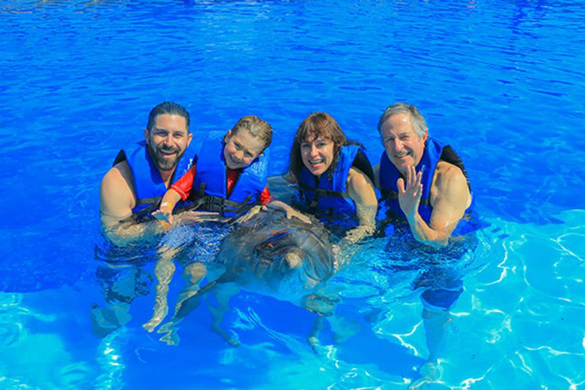 Cuatro personas con chalecos salvavidas azules sonriendo y posando con un delfín en una piscina con agua azul clara
