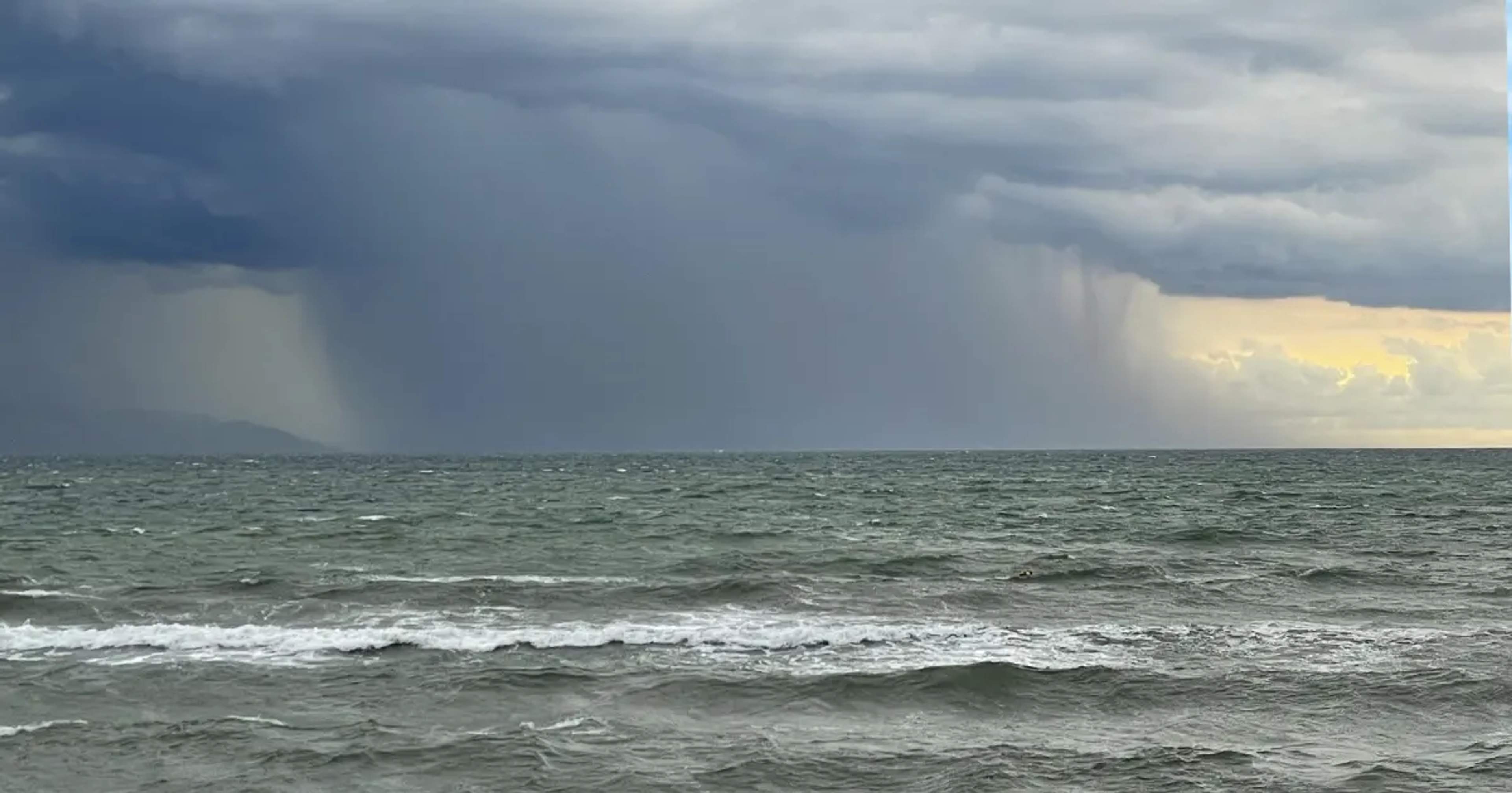Dark storm clouds over the ocean, with rainfall in the distance and a small break of sunlight near the horizon.