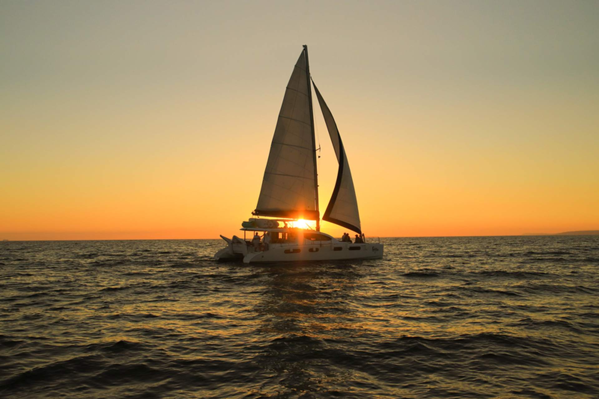 A sailboat glides across the ocean at sunset, with the sun setting behind the sails, creating a picturesque scene.