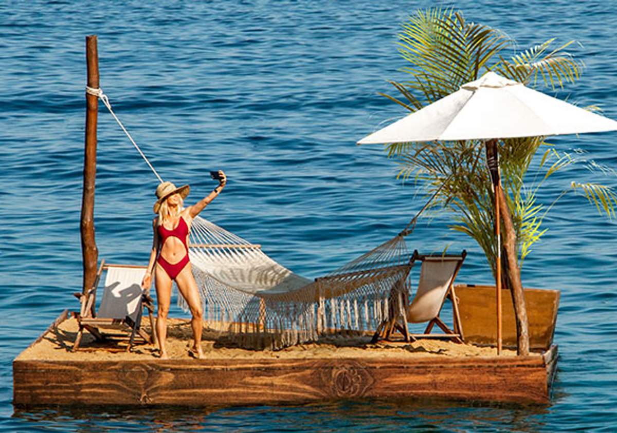 Woman in a red bikini taking a selfie on a small floating platform with a hammock, umbrella, and chairs on the ocean.