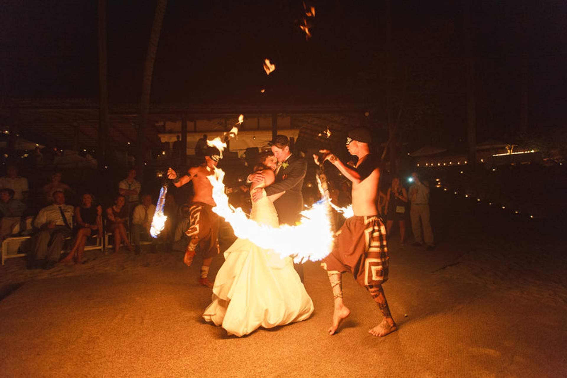 Una novia y un novio disfrutan de una actuación de danza con fuego en su boda nocturna en la playa, rodeados de invitados.