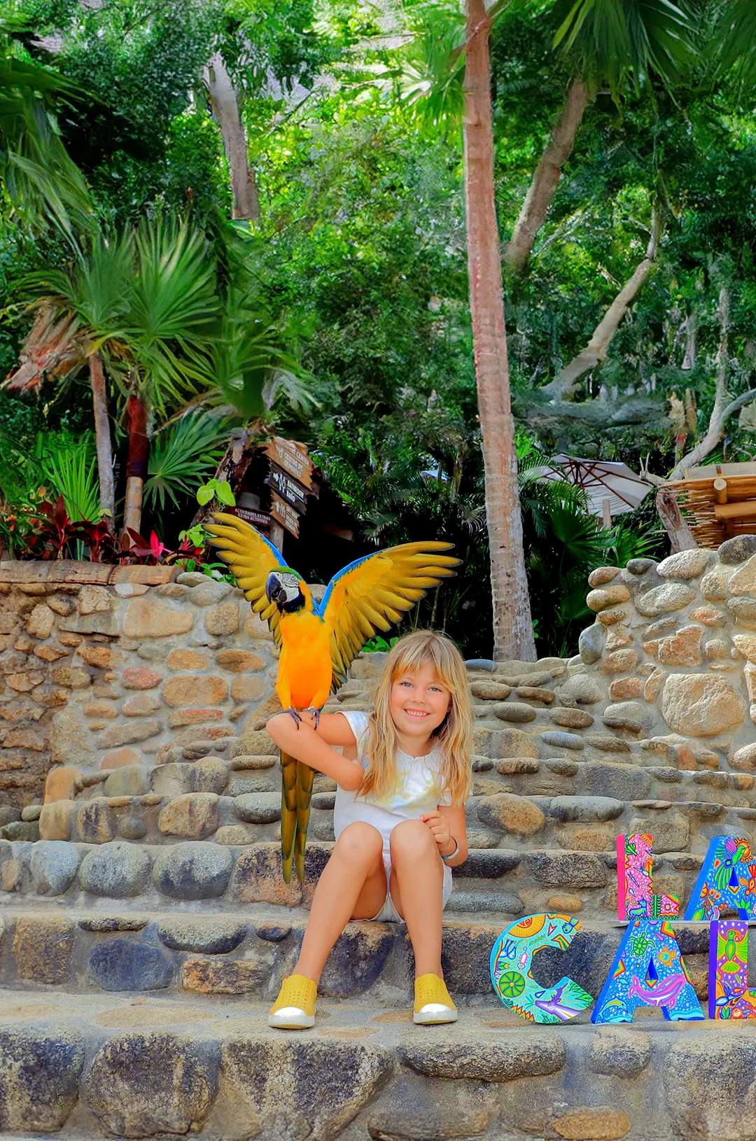 Smiling girl with a macaw on her arm, sitting by "Las Caletas" sign.