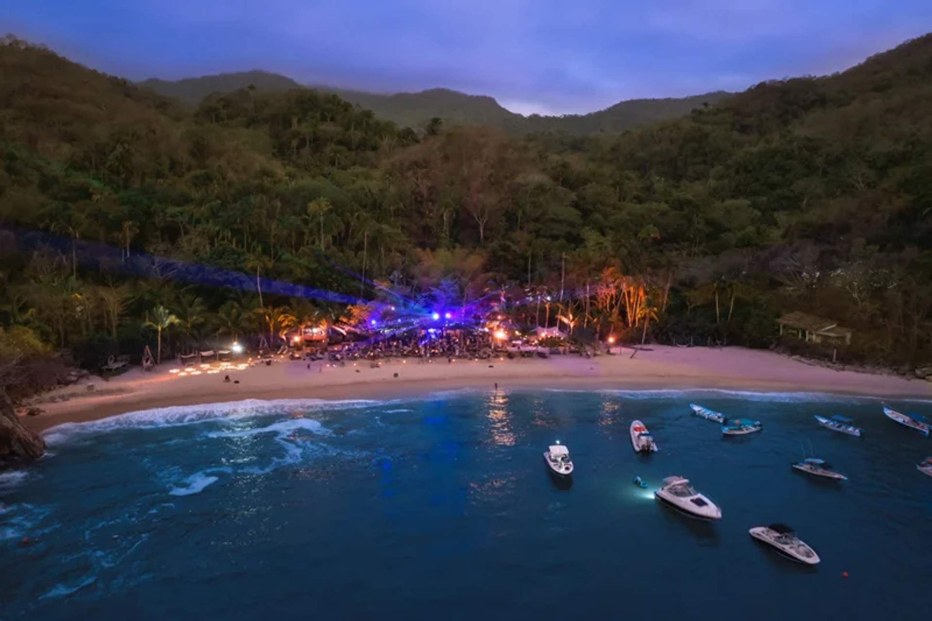 An aerial view of a tropical beach at dusk, with boats anchored near the shore. Festive lights and a gathering of people create a lively atmosphere, surrounded by lush greenery and hills in the background.