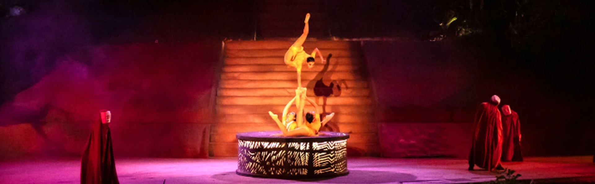Acrobats performing a balancing act on a circular platform, illuminated by dramatic lighting with steps in the background. Two figures in red robes are watching the performance from the side 