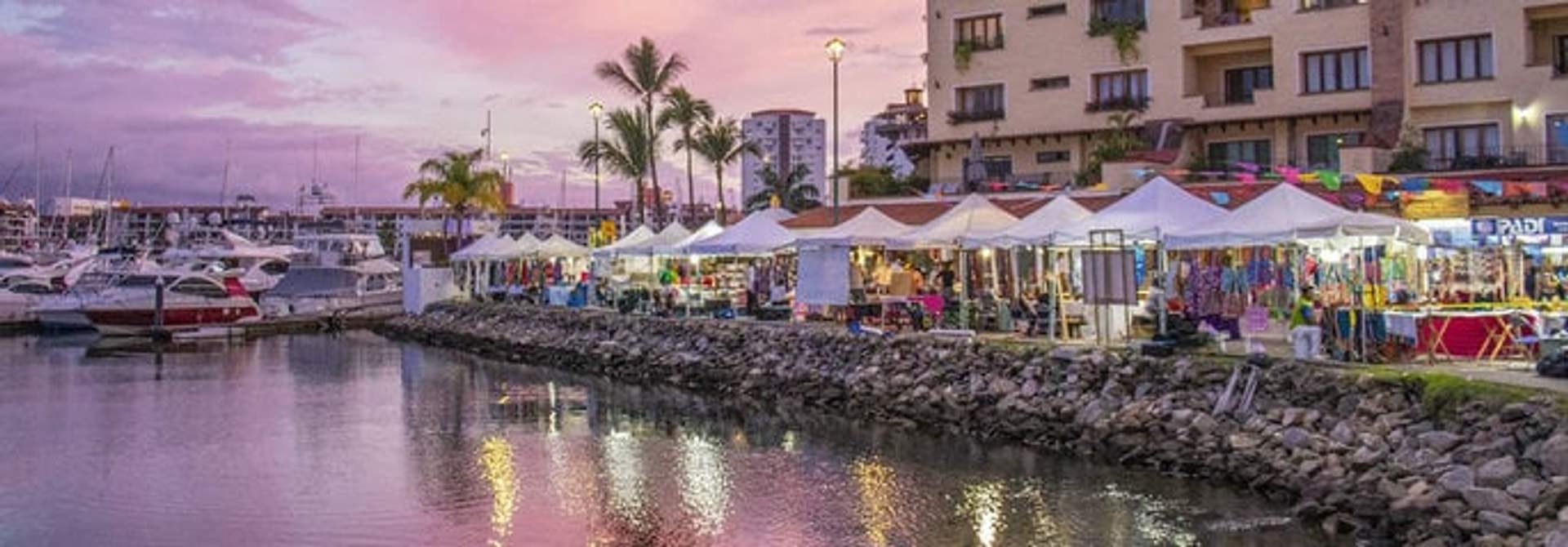 A bustling market with white tents set up along the waterfront at Marina Vallarta, with boats docked and a sunset sky.
