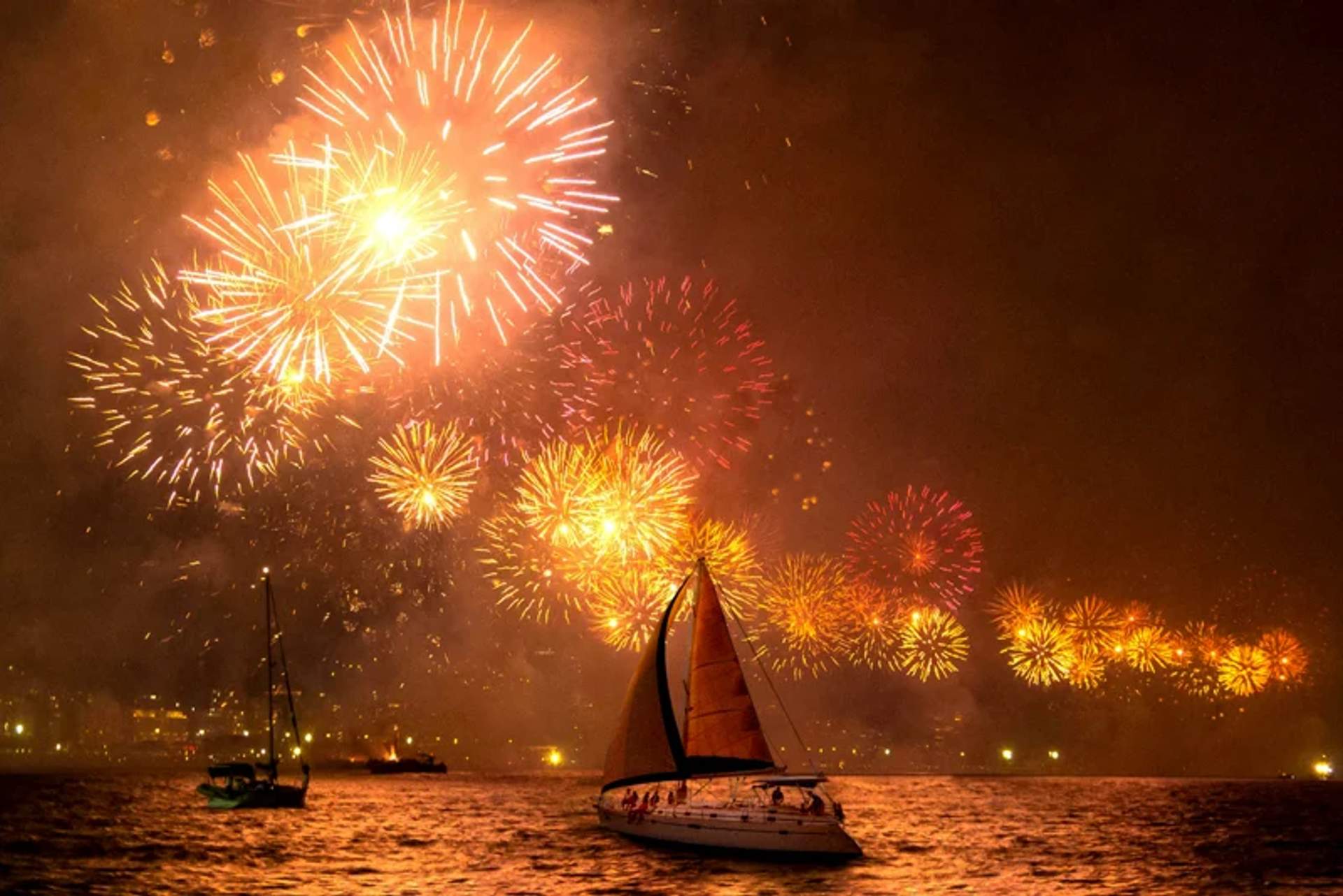 Un espectacular espectáculo de fuegos artificiales iluminando el cielo nocturno sobre el océano, con un velero en primer plano. Las explosiones vibrantes en tonos de naranja y amarillo crean una escena festiva y de celebración.