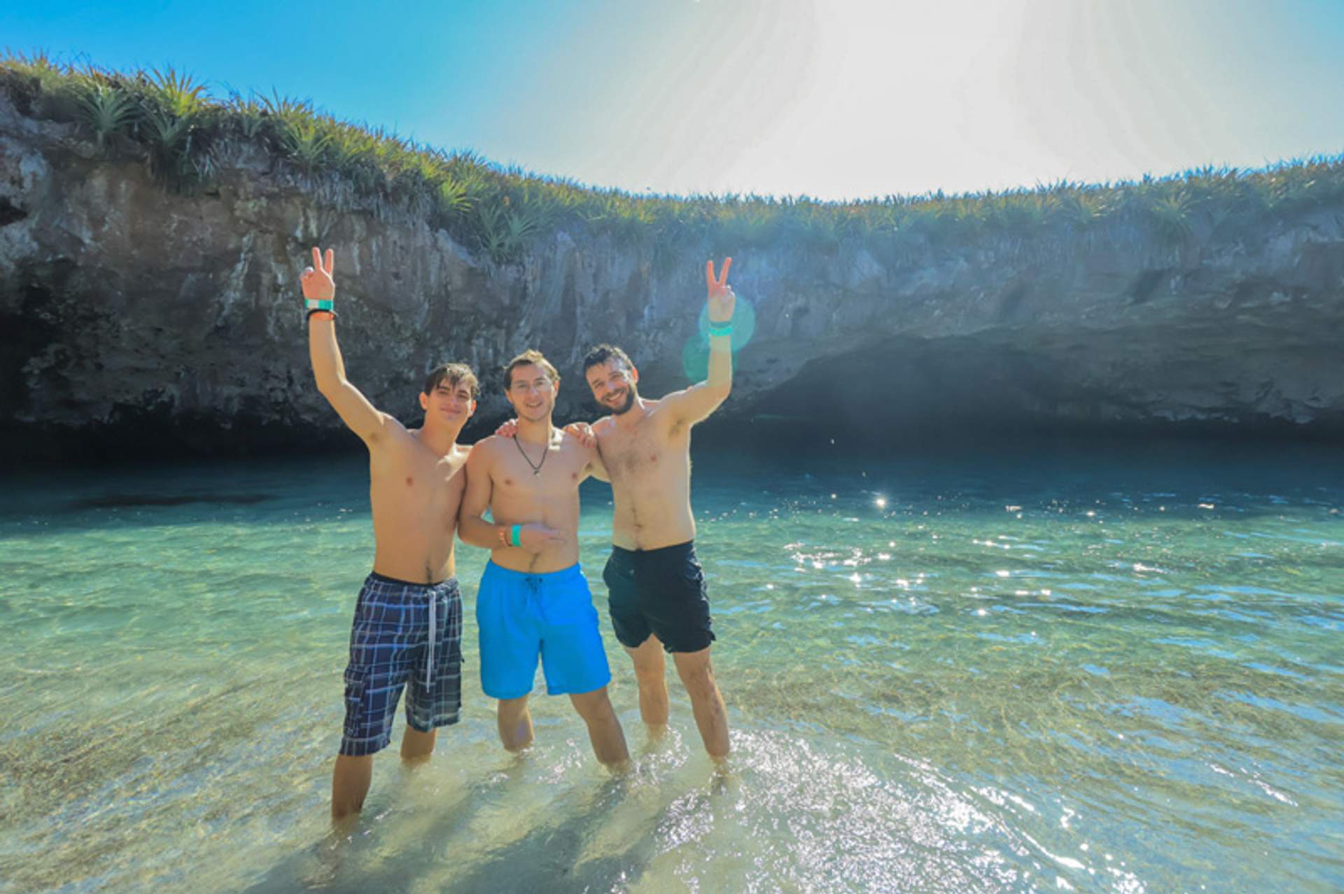 Tres hombres posando y haciendo signos de paz en agua poco profunda con una cueva rocosa al fondo.
