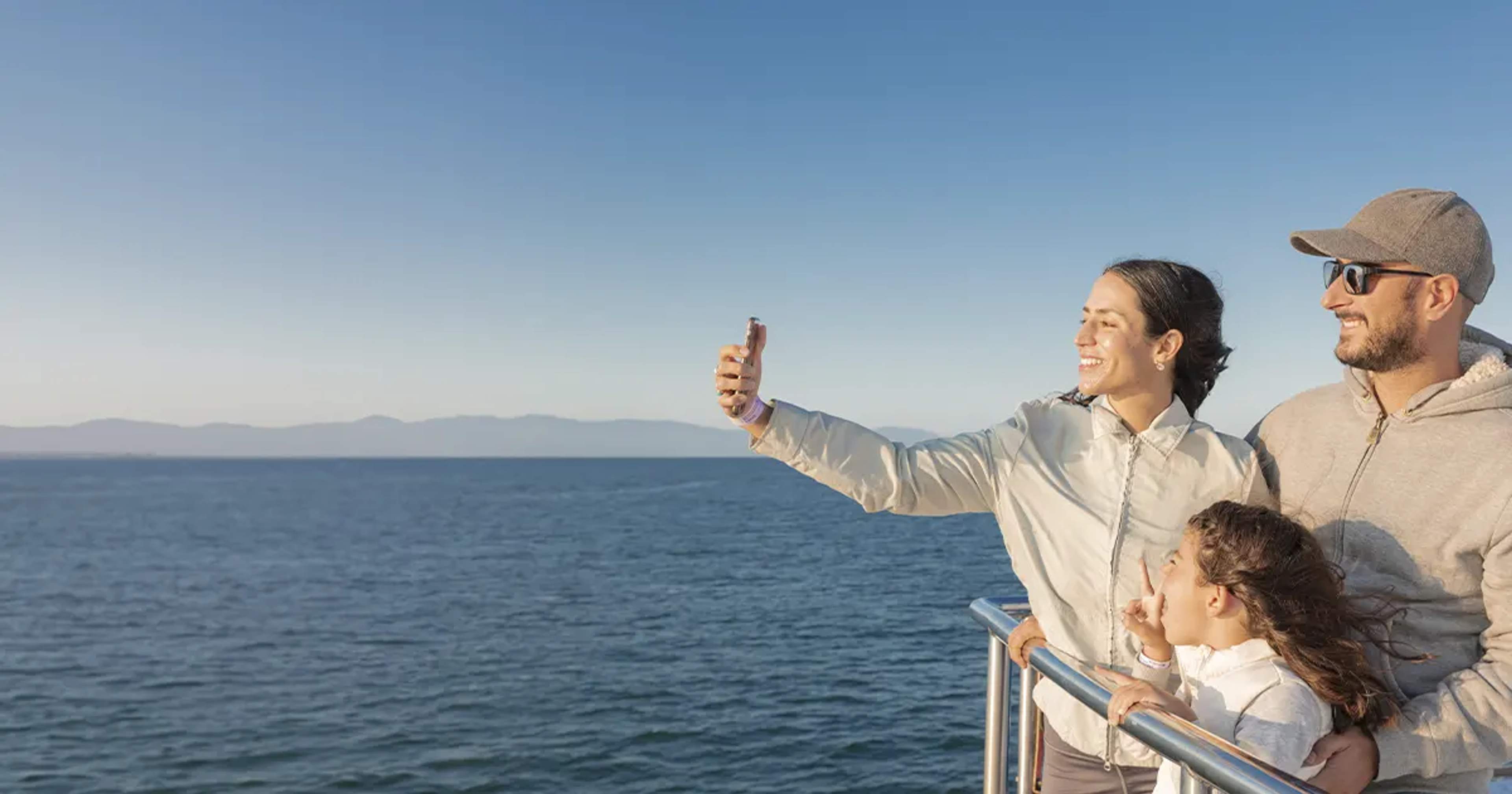 Una familia tomándose una selfie en un barco, con el océano y montañas distantes al fondo bajo un cielo azul despejado.