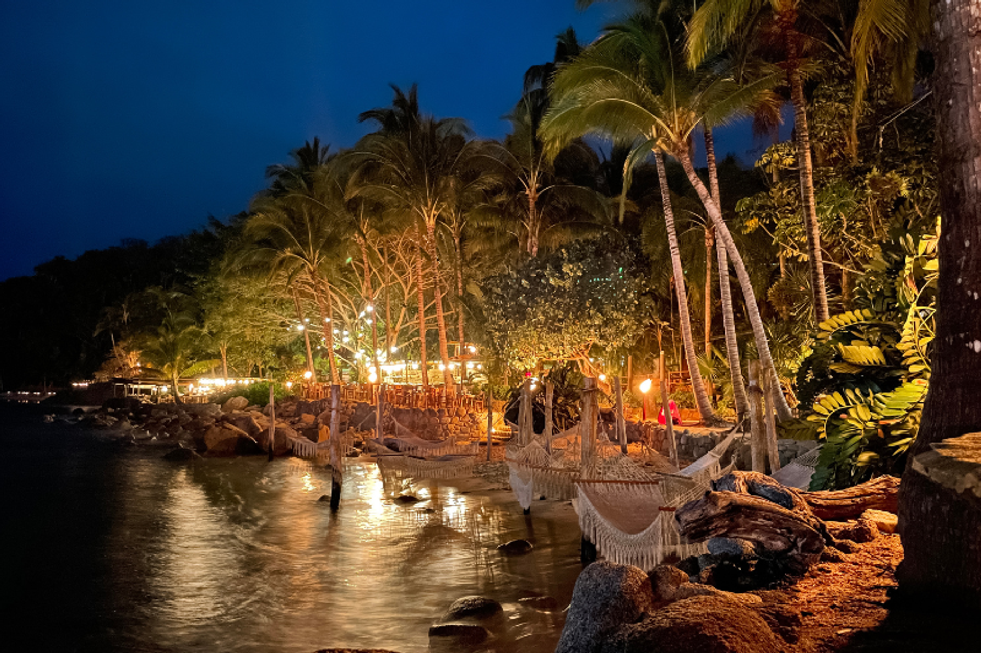 A tropical beach scene at night, illuminated by warm lights among the palm trees. Hammocks are strung between the trees, and the water gently laps against the rocky shore, creating a serene atmosphere.
