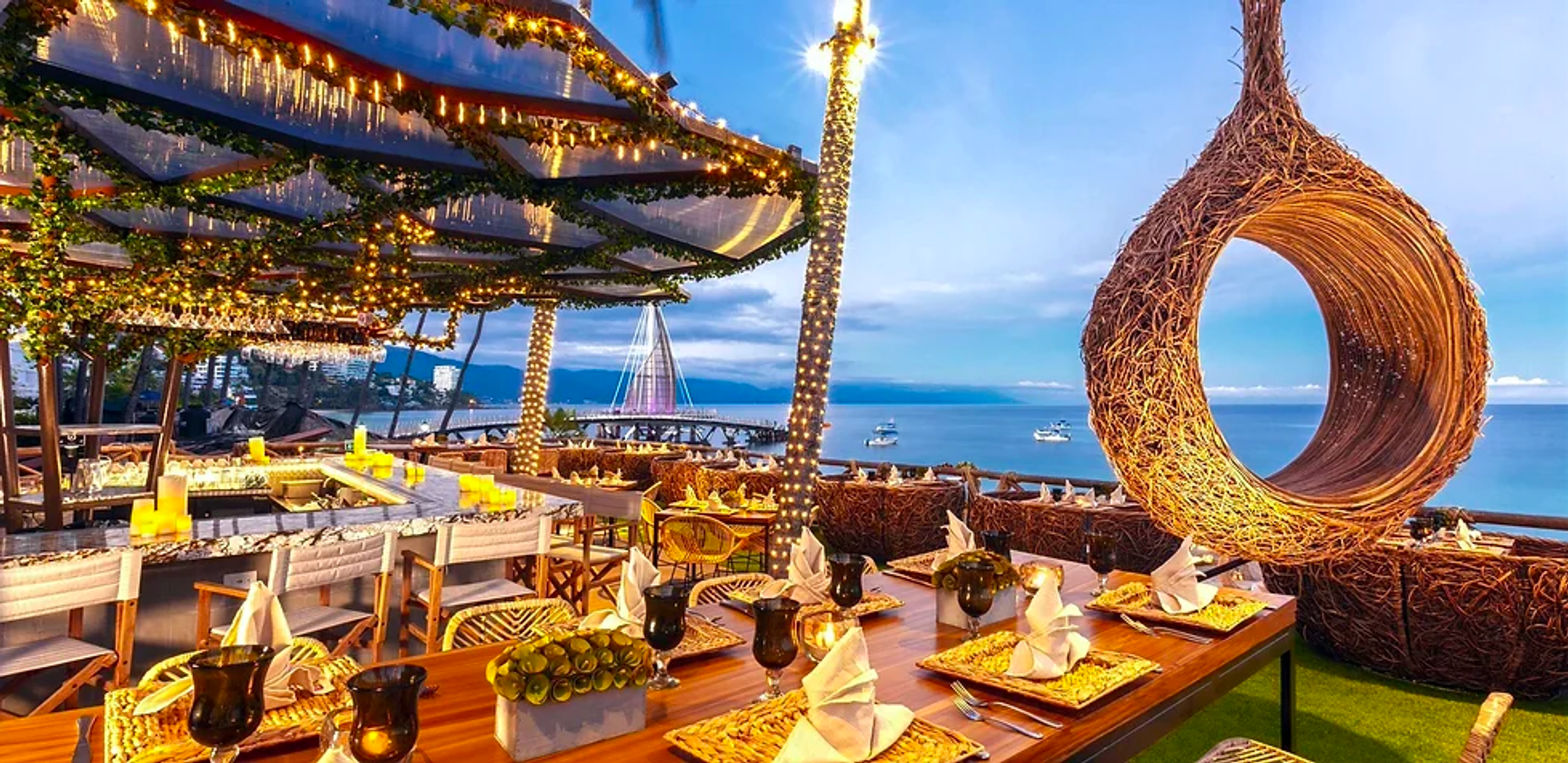 Restaurante al aire libre decorado con luces y adornos, con mesas elegantemente preparadas y vistas al mar al atardecer.