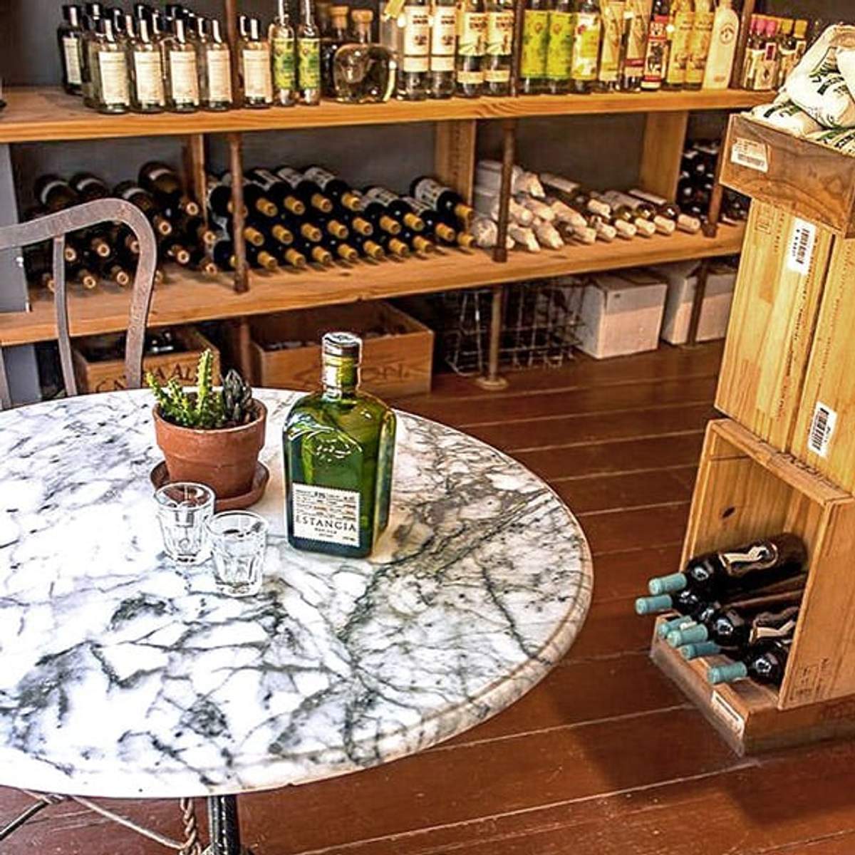 Mesa de mármol con una botella de mezcal y vasos de chupito en una tienda de vinos, rodeada de estantes con botellas de vino.