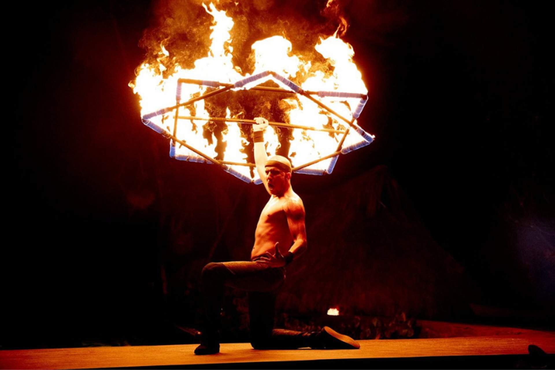 Un artista sin camisa arrodillado sobre una rodilla mientras sostiene un gran objeto de fuego en forma de estrella sobre su cabeza, con un fondo oscuro.