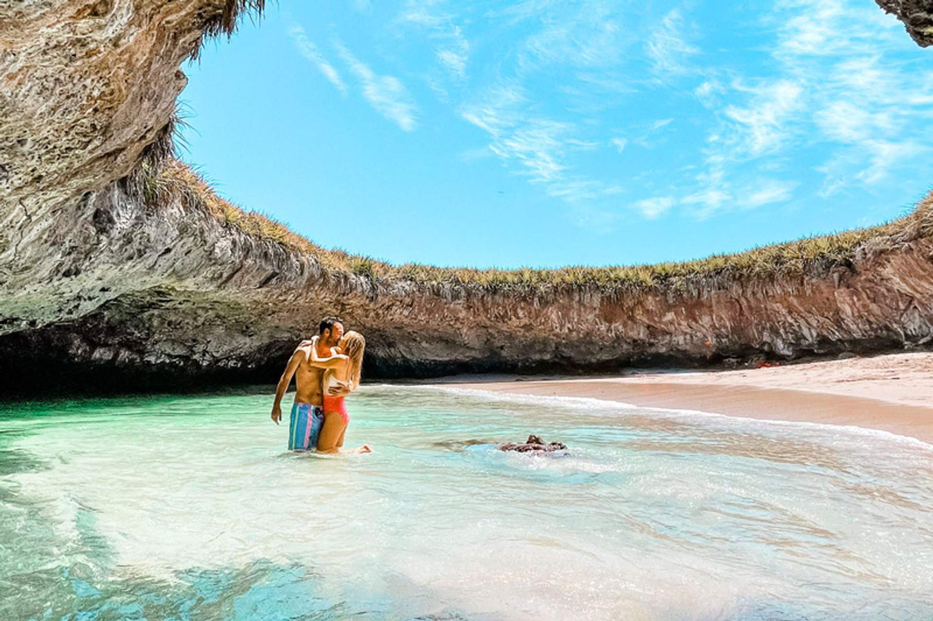 Pareja abrazándose en agua poco profunda bajo un arco natural de roca con vegetación.