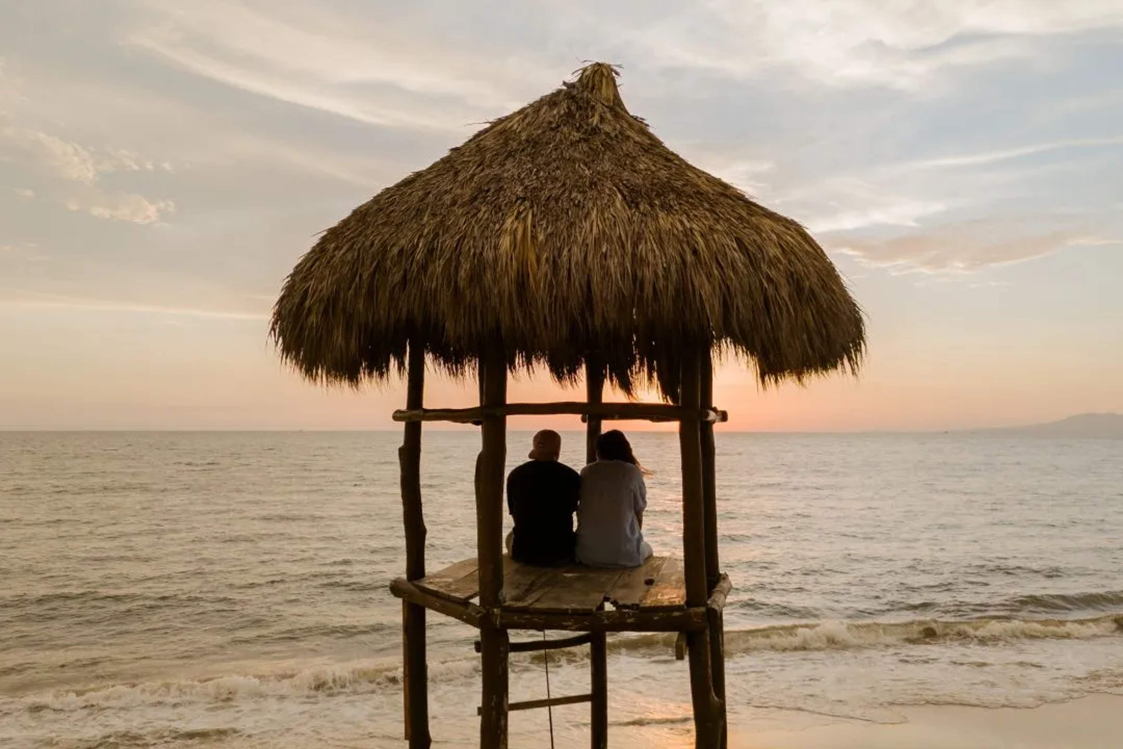 Dos personas se sientan bajo una rústica palapa de madera con vista al océano durante un atardecer sereno en Puerto Vallarta.