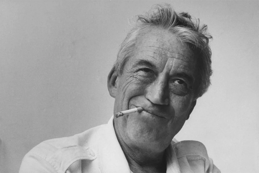 John Huston, sonriente con un cigarro, refleja un instante espontáneo de encanto y rebeldía.