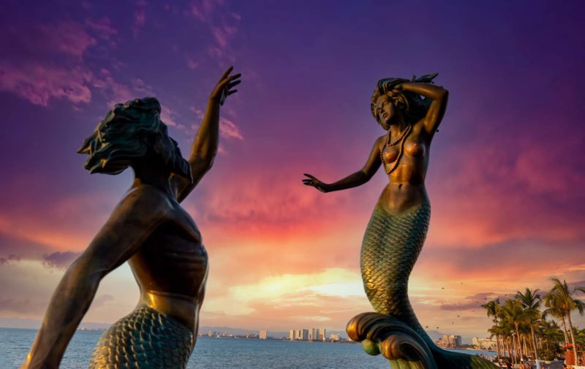 A stunning view of two mermaid statues on the Malecon boardwalk in Puerto Vallarta, Mexico, during sunset. The statues are highlighted against a dramatic sky with shades of purple, pink, and orange, and the city skyline is visible in the distance.