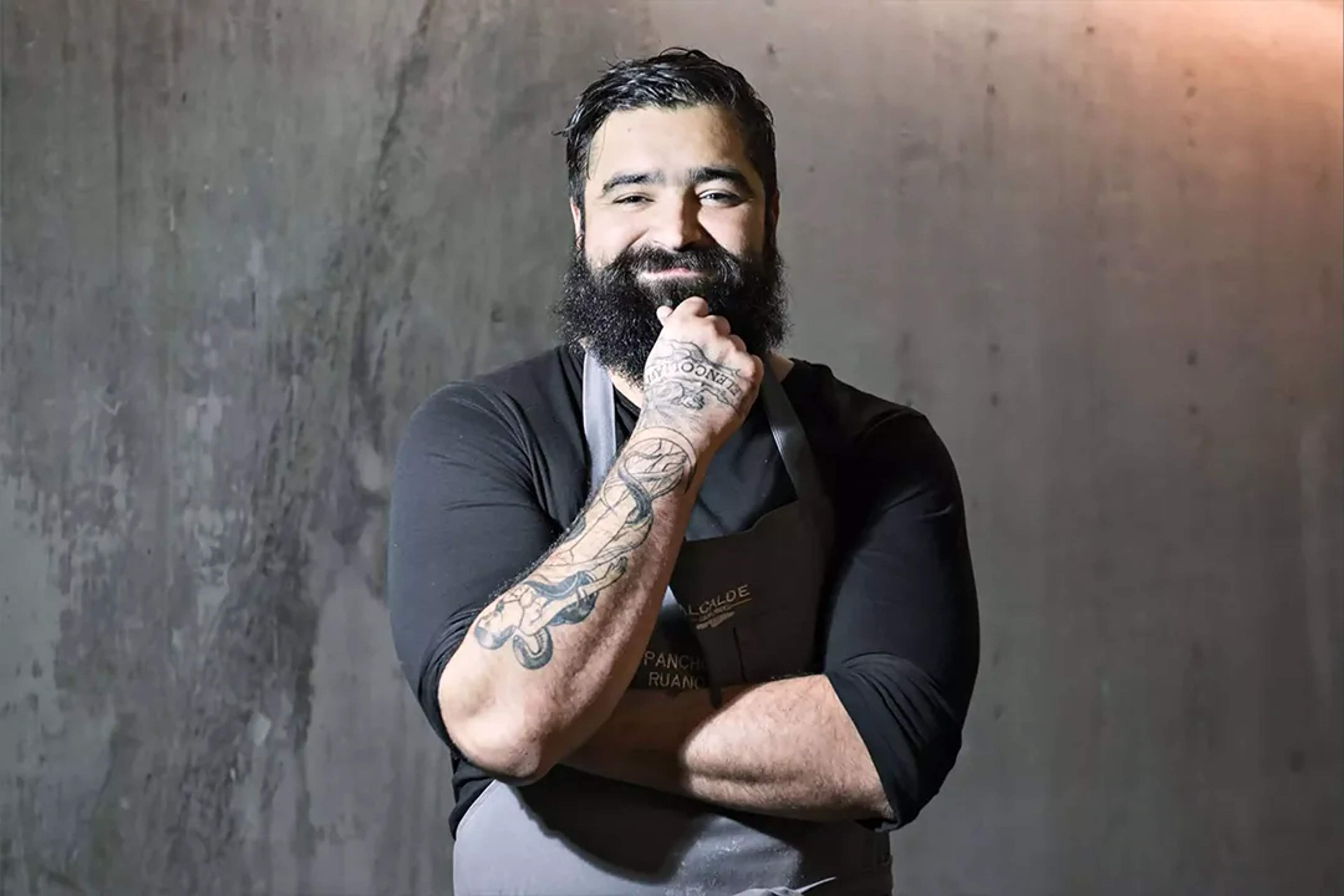 Retrato del chef Paco Ruano con mandil, sonriendo con confianza y brazos tatuados cruzados.