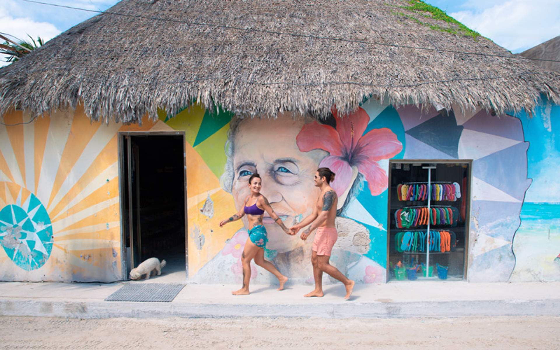 Pareja de la mano frente a un mural colorido en un edificio con techo de paja.