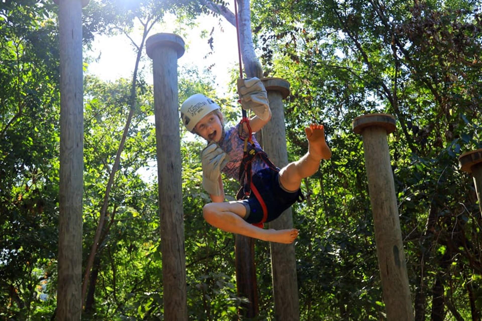 Un niño sonriente disfruta de una aventura en tirolesa, equipado con casco y guantes, rodeado de árboles y pilares de madera.