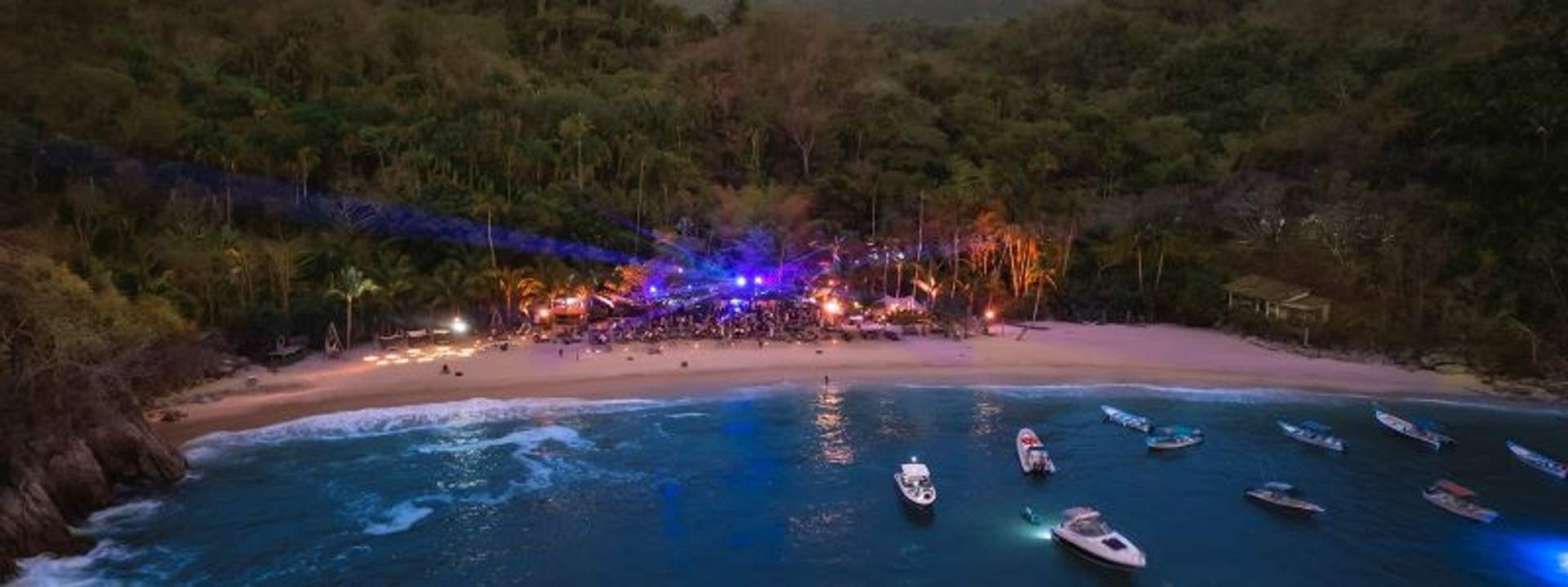 Aerial view of a lively beach party at night in a secluded cove surrounded by dense greenery. The beach is illuminated with colorful lights and music, while several boats are anchored near the shore.