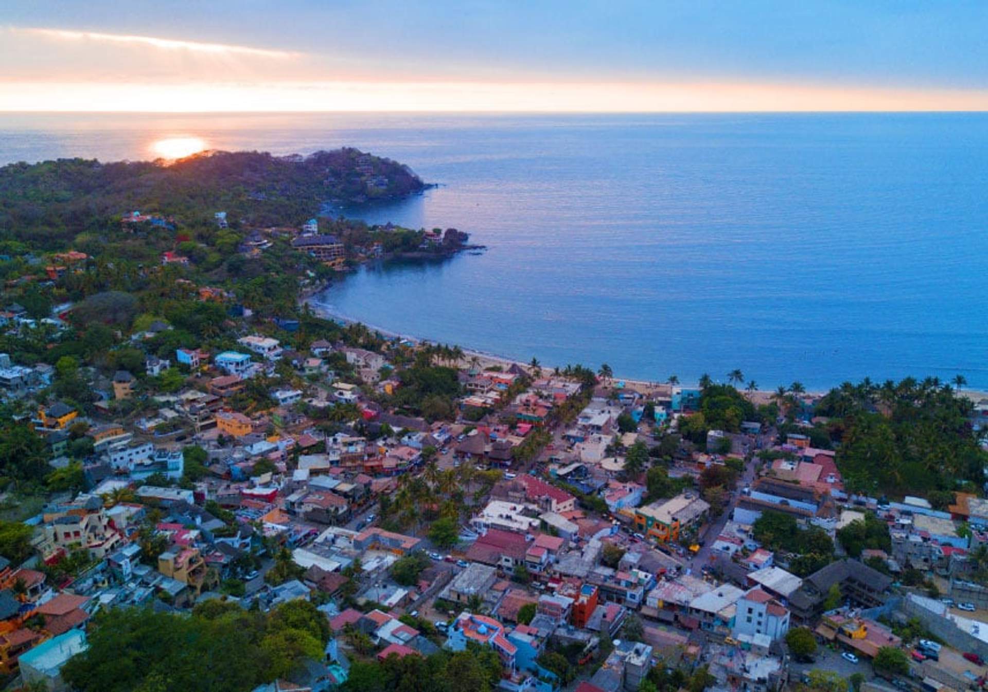 Aerial view of Sayulita, a coastal town in Mexico, with colorful buildings, lush greenery, and a serene ocean at sunset.