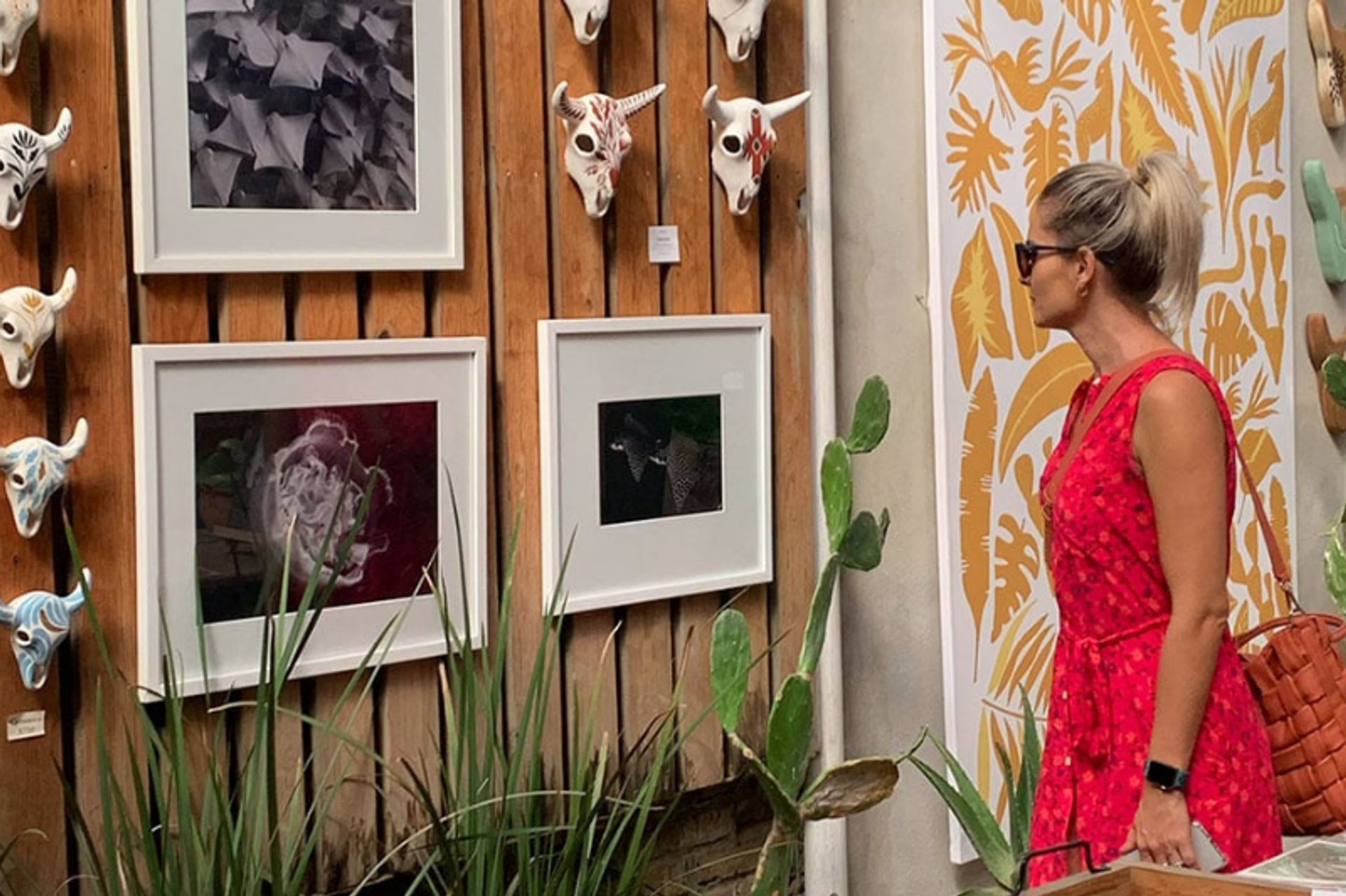 A woman in a red dress and sunglasses looking at artwork displayed on a wooden wall. The display includes framed photographs and decorative skull masks, with plants adding to the aesthetic.