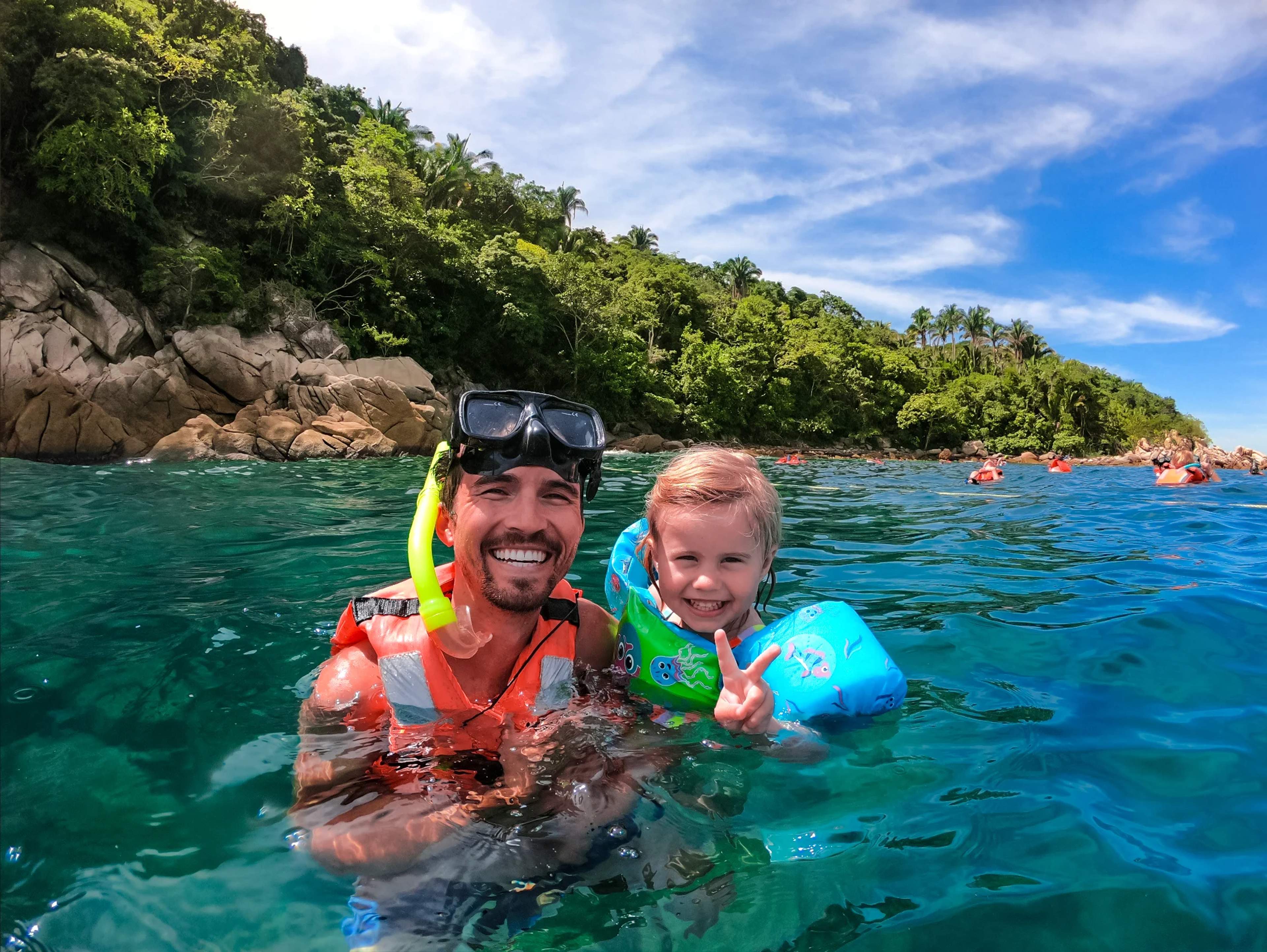 A man and a child in snorkeling gear smile and pose in the ocean, with a lush green shoreline and other swimmers in the background.