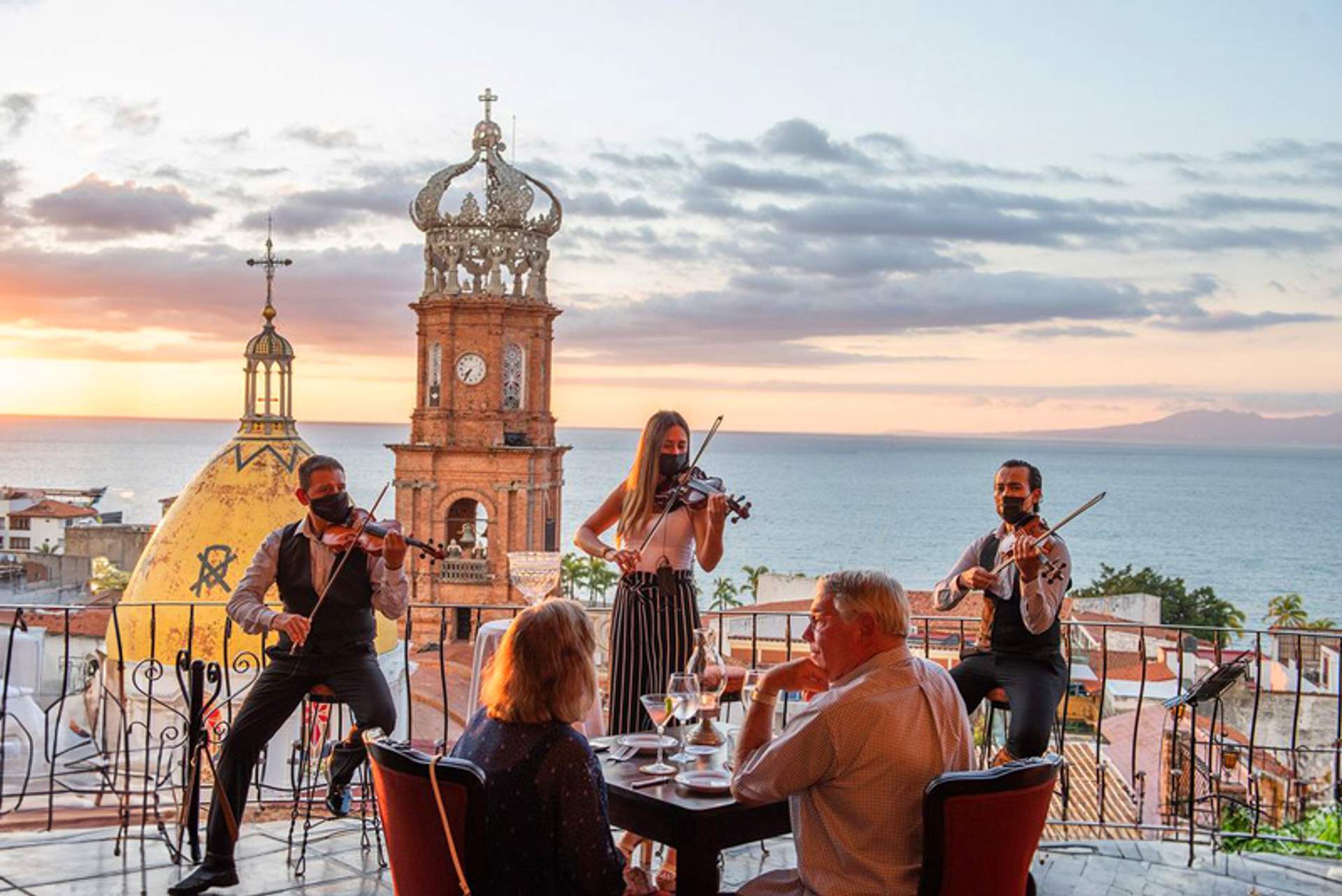 Un grupo de personas cenando al aire libre con vista de la iglesia en Puerto Vallarta y el mar al atardecer, acompañados de músicos tocando violines.