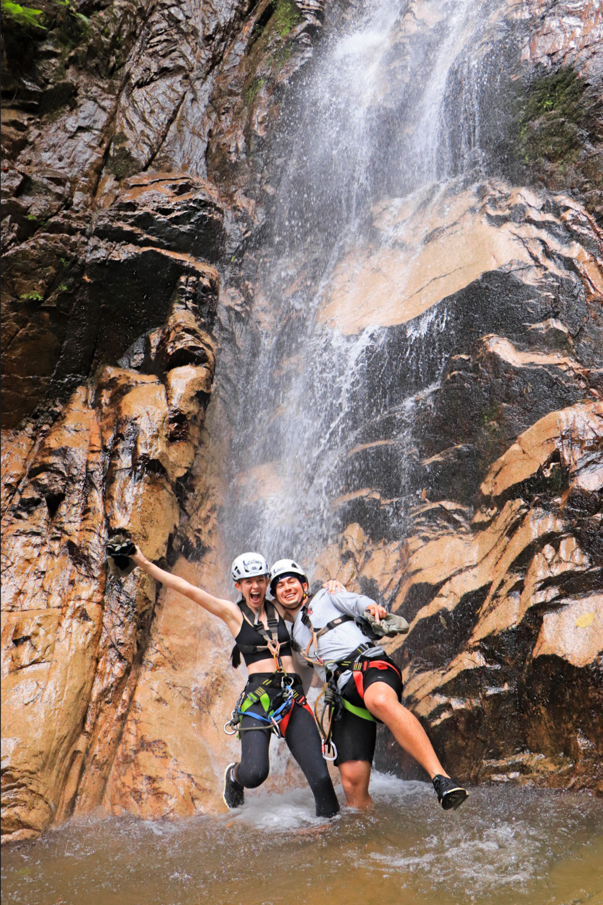 Dos personas con cascos y arneses de pie alegremente debajo de una cascada con rocas al fondo. Están sonriendo y posando, con una persona levantando el brazo con entusiasmo.