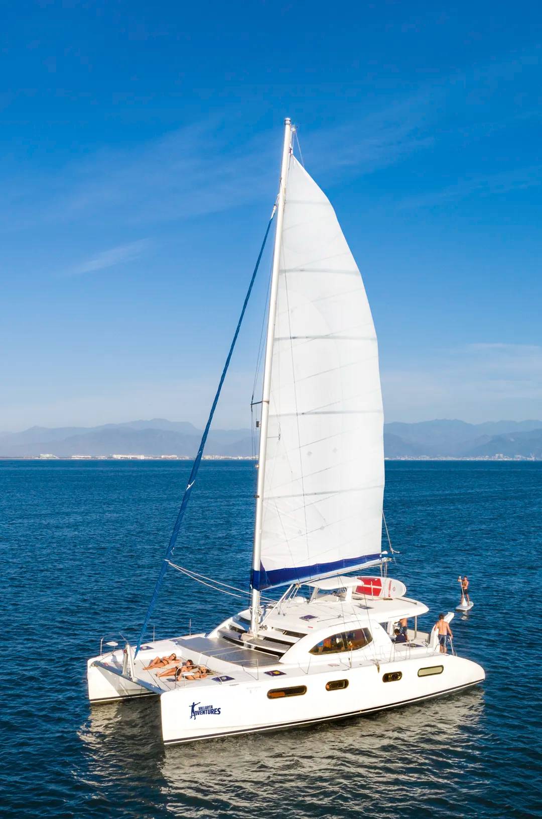 A catamaran sailing in Puerto Vallarta's blue waters, with people relaxing and paddleboarding nearby.