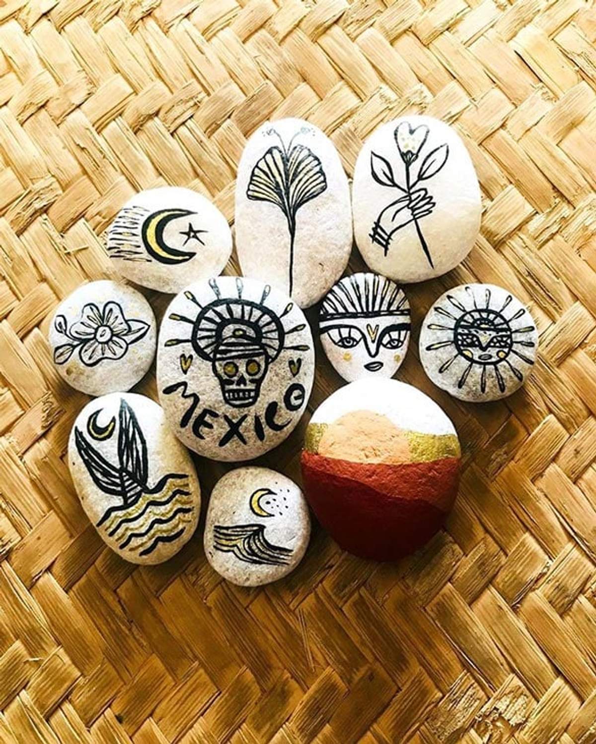 Piedras pintadas a mano con varios diseños, incluyendo "México," flores y caras, dispuestas sobre una estera tejida.