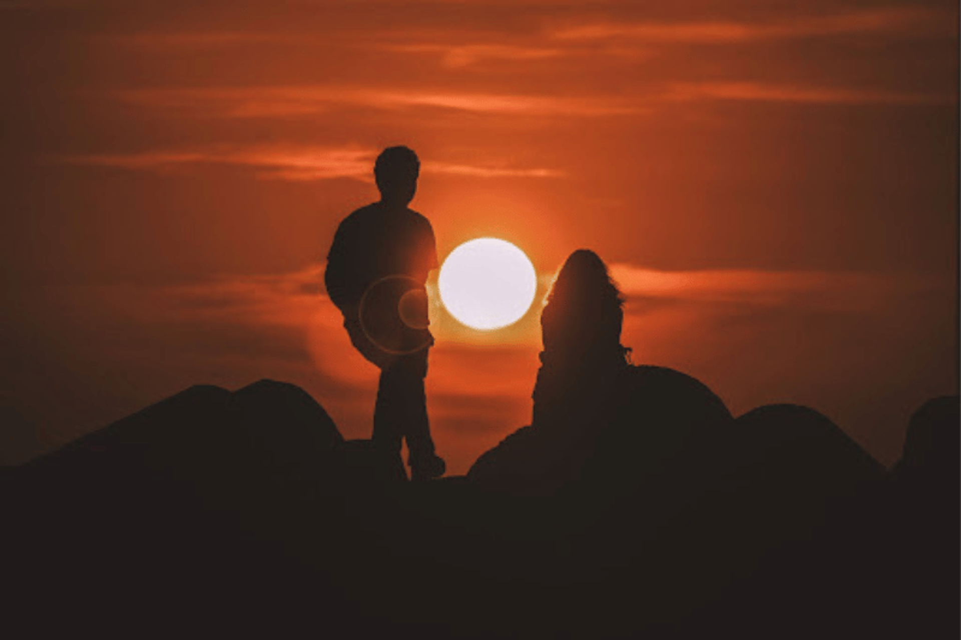 A silhouette of a couple against a vibrant orange sunset, standing on rocky terrain. The sun is positioned low in the sky, creating a dramatic and romantic scene.