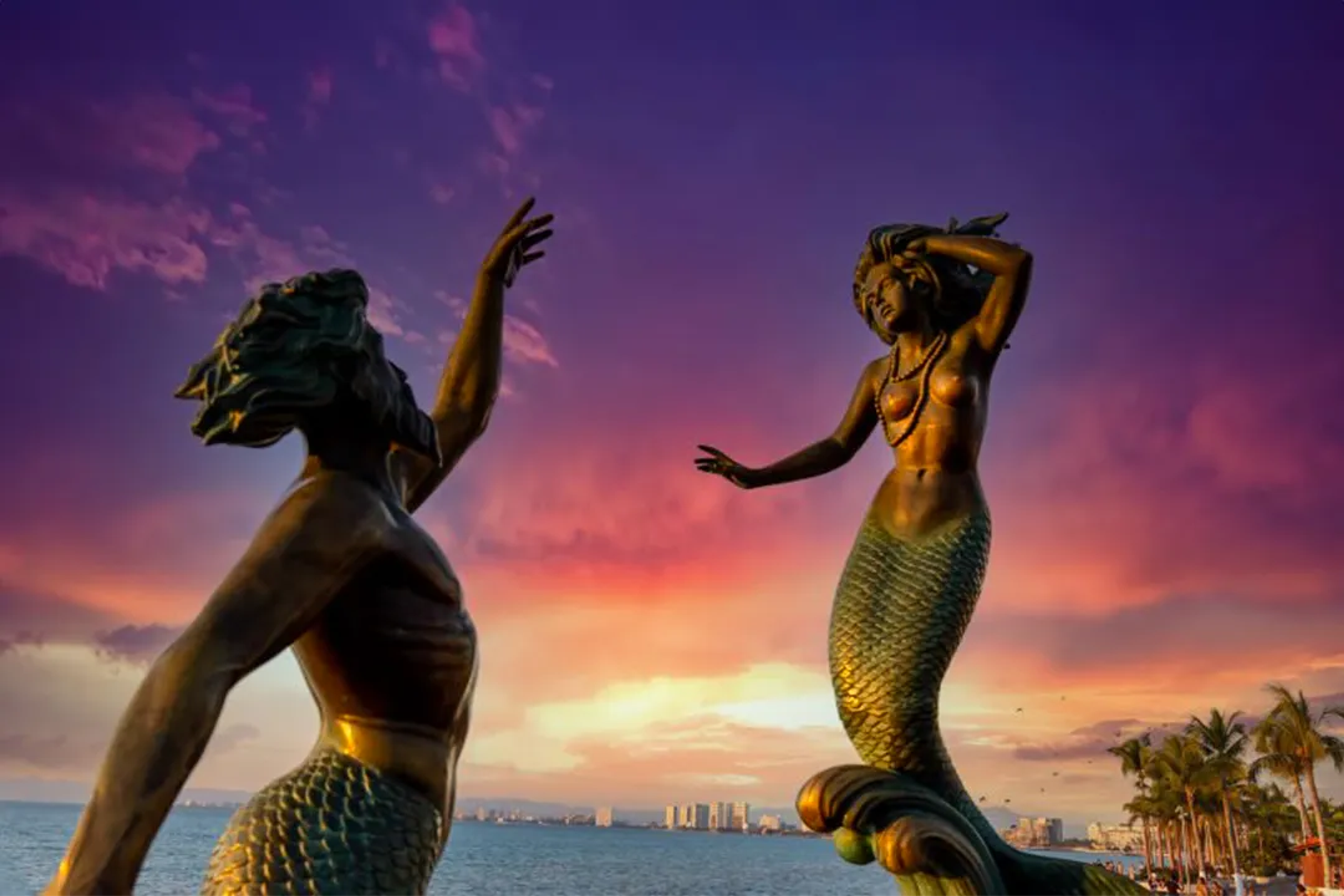 Mermaid sculptures on the Puerto Vallarta Malecón at sunset, with a vibrant sky.