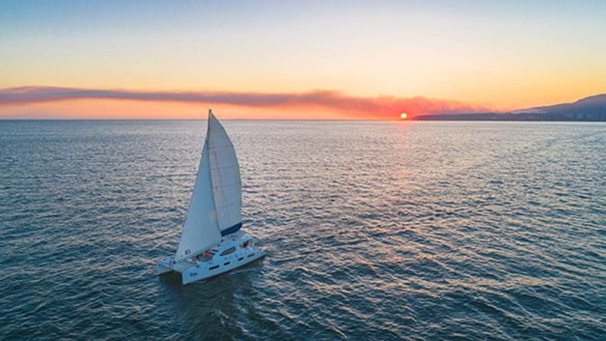 A sailboat on the open sea at sunset, with the sun setting near the horizon and calm waters surrounding it.