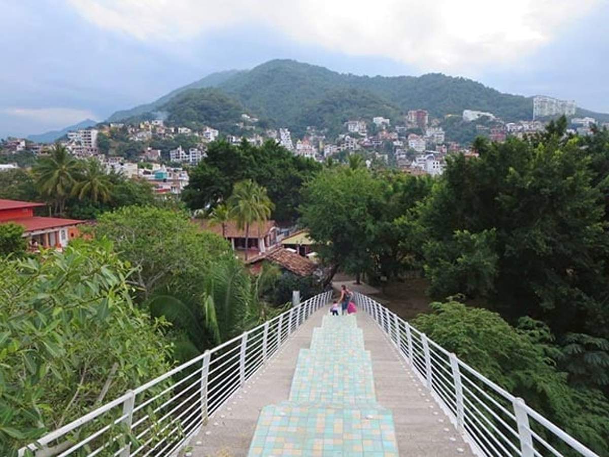 Puente peatonal desde Gringo Gulch que conduce a un entorno verde con el paisaje urbano y las montañas de Puerto Vallarta al fondo.