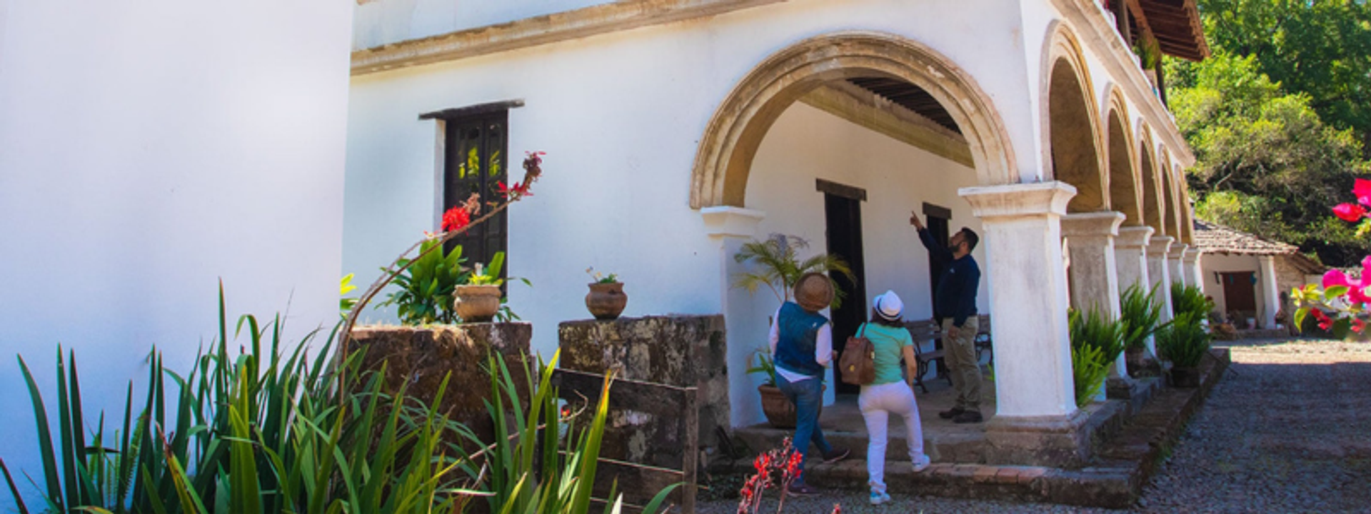 Three people exploring an old white colonial building with arched doorways and stone steps. The building is surrounded by greenery and potted plants, creating a historical and serene atmosphere.