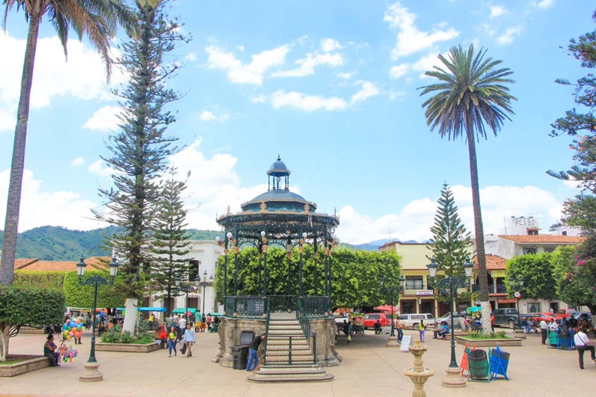 Town square with a central gazebo, tall palm trees, and people strolling under a bright blue sky. / Plaza del pueblo con un quiosco central, altas palmeras y personas paseando bajo un cielo azul brillante.