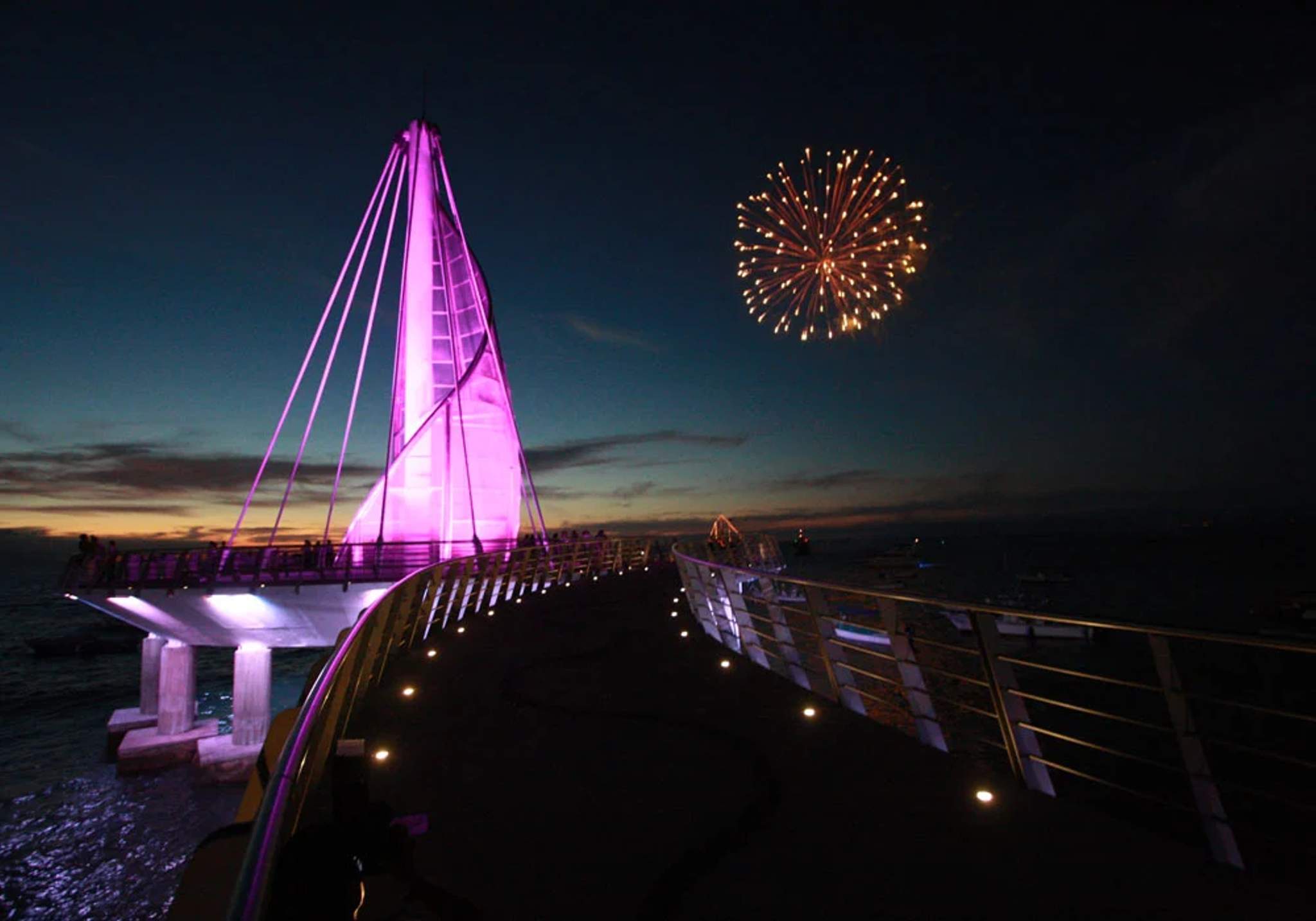 The Los Muertos Pier in Puerto Vallarta illuminated with pink lights at night, with fireworks exploding in the sky above. The modern sail-shaped structure stands out against the dark evening sky in Puerto Vallarta.