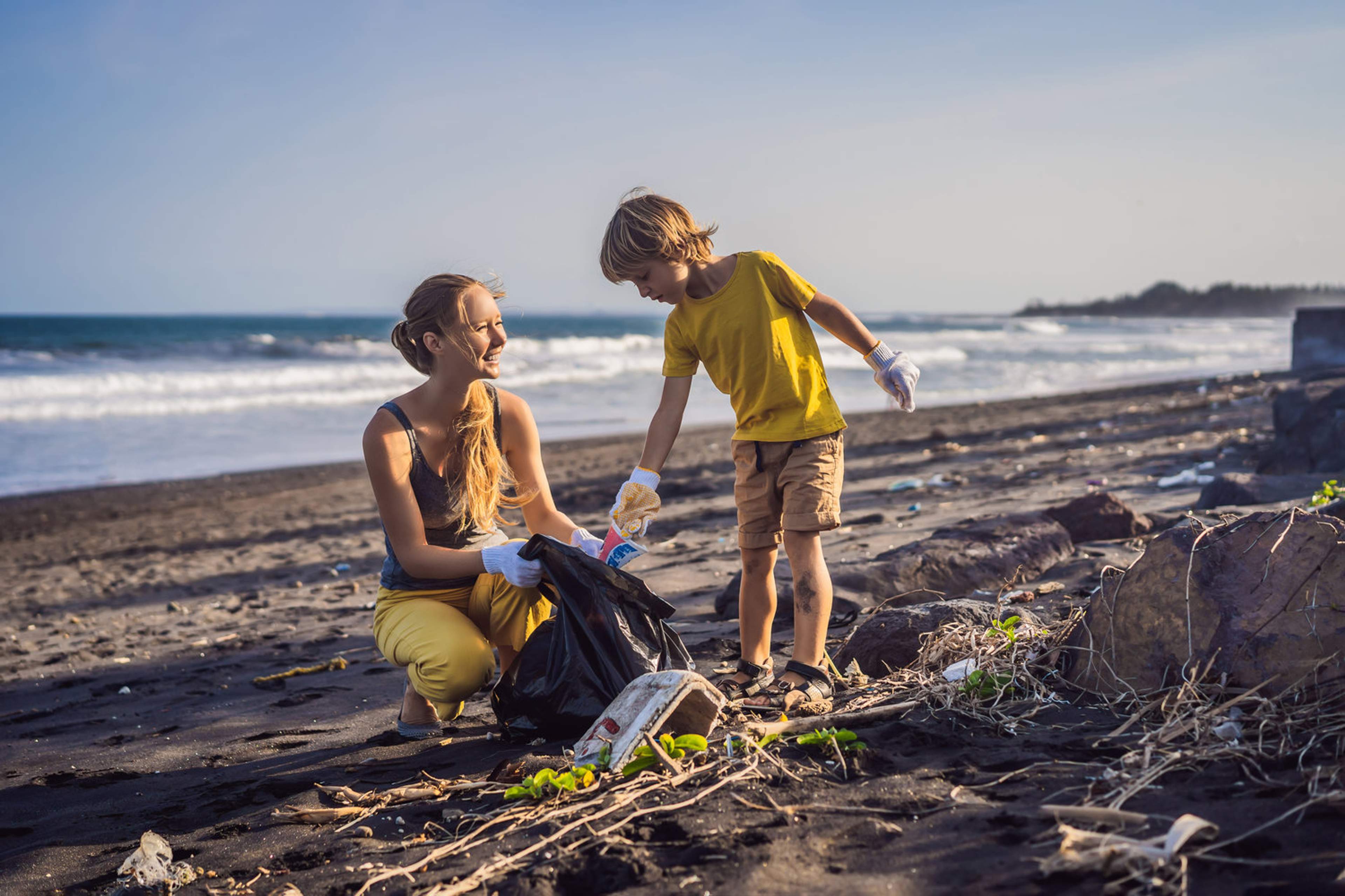 A woman and child wearing gloves collect trash on a beach, participating in a cleanup effort to protect the environment.