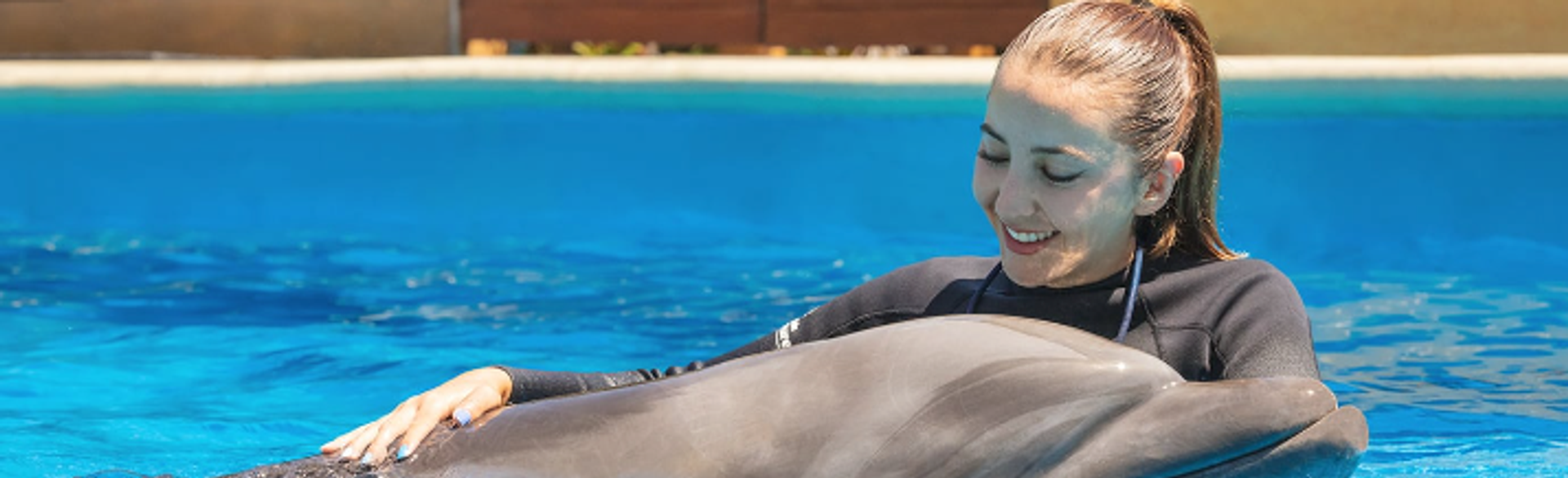 A woman smiling while hugging a dolphin in a pool. She is wearing a wetsuit, and both appear to be enjoying the interaction.