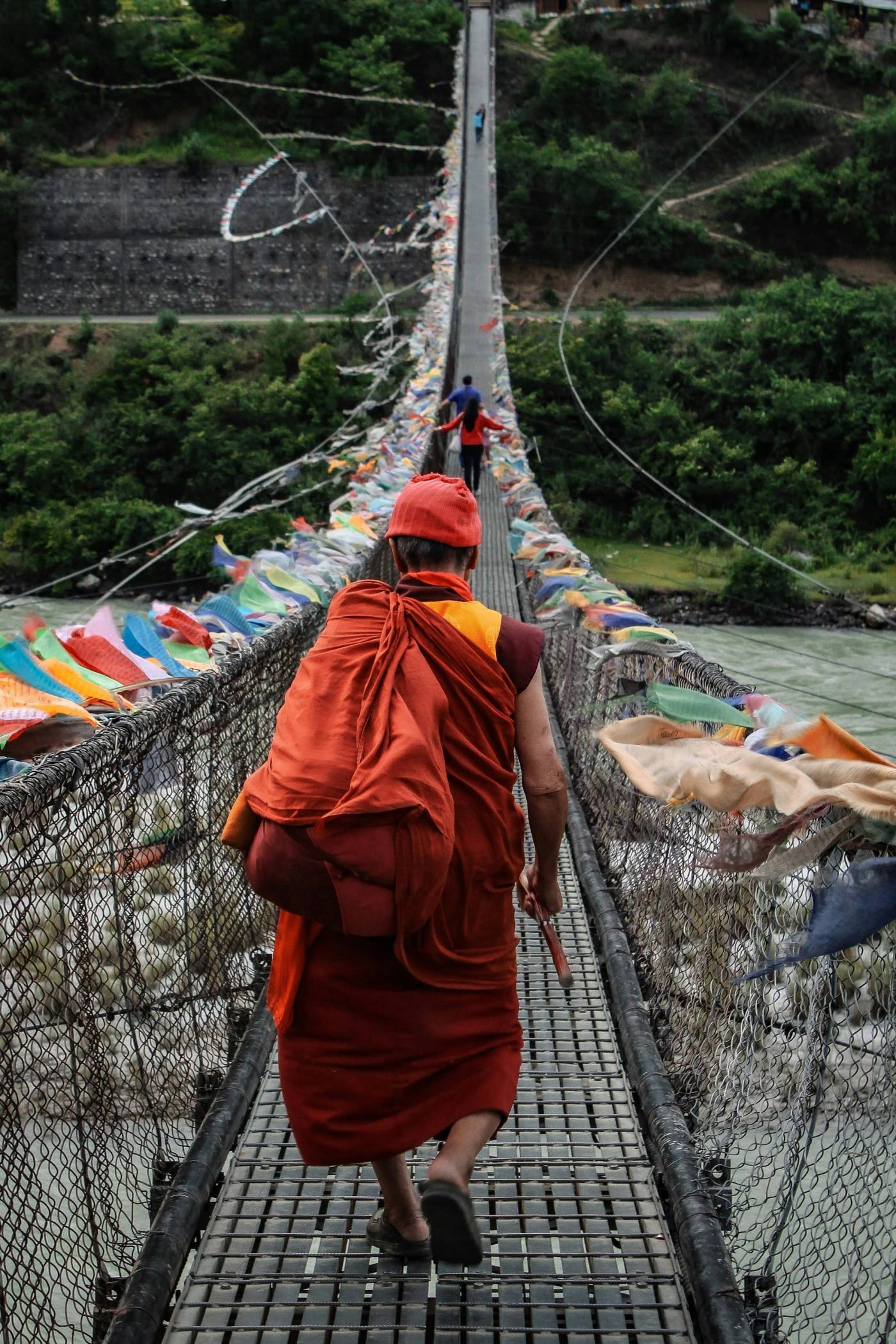 A monk in red robes crosses a long suspension bridge hung with prayer flags