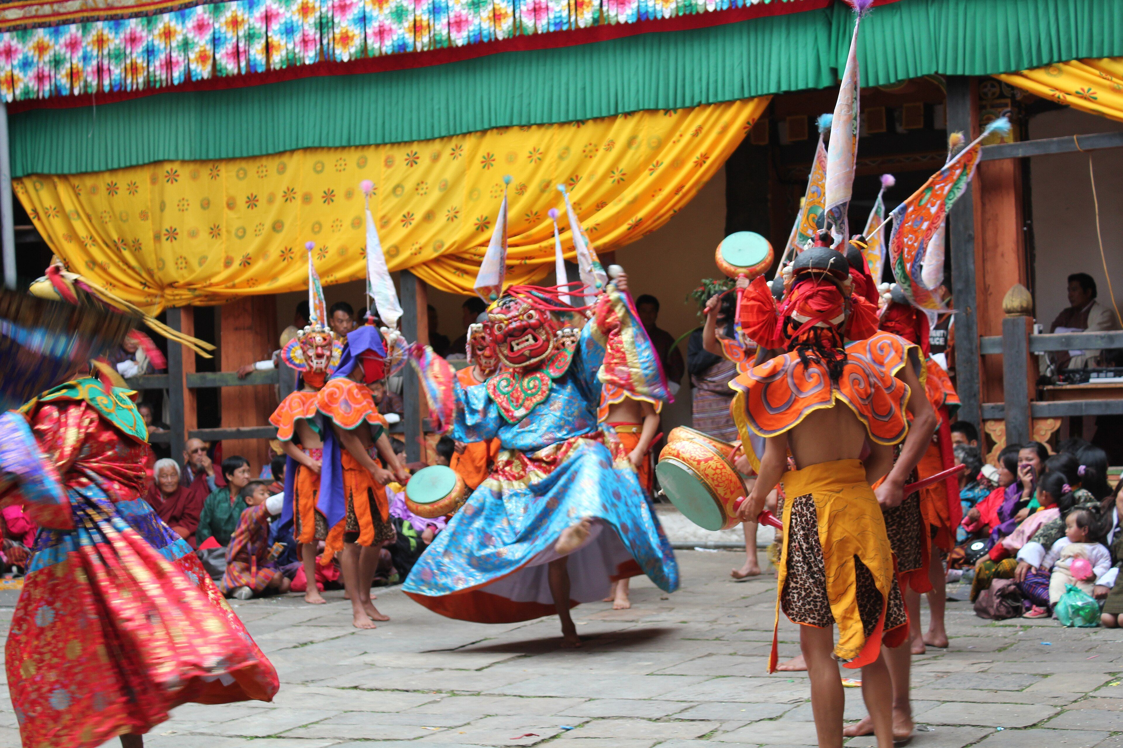 Masked dancers in a dzong courtyard: one in a flowing blue brocade robe and red mask spins mid-step while others in orange collars and leopard-print skirts beat hand drums, with families seated under yellow drapes