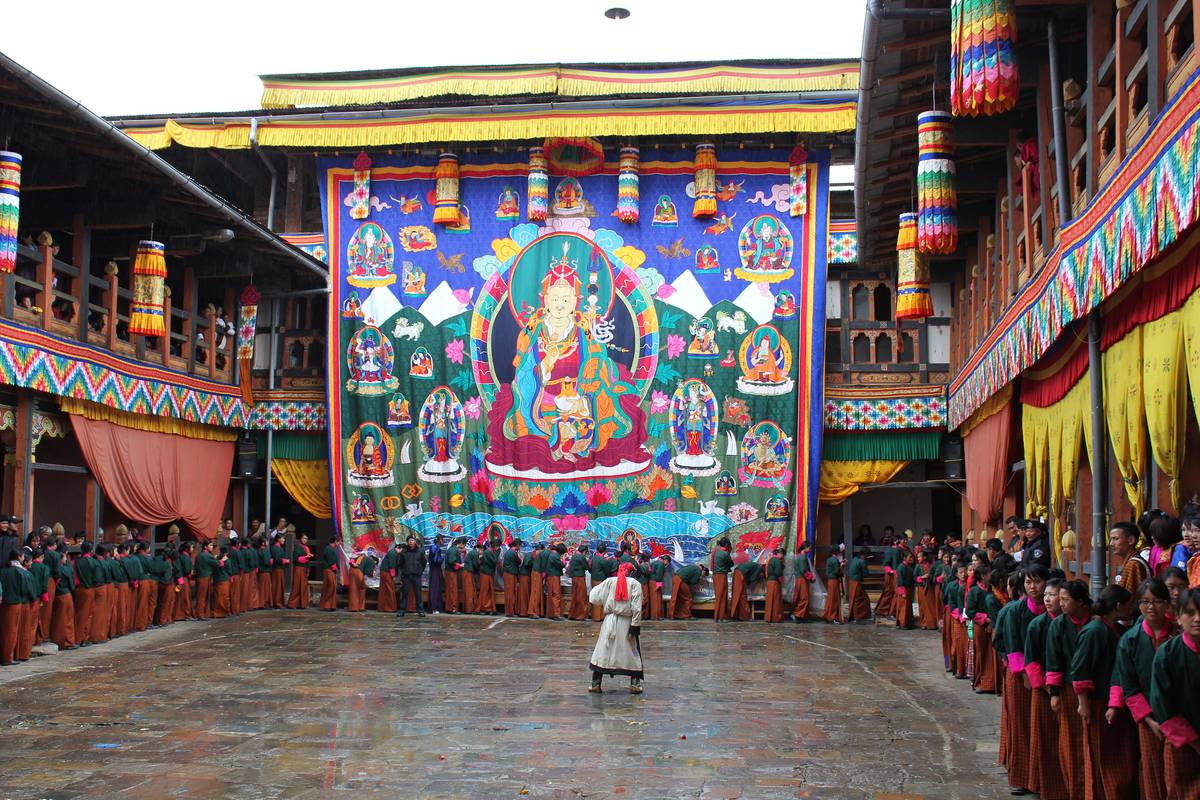 A giant appliqué image of Guru Rinpoche hangs across one side of a dzong courtyard while lines of schoolgirls in green jackets and striped kira file past it on a wet stone floor