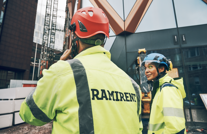 En man som pratar i mobiltelefon ute på en byggarbetsplats och en kvinna som ler och tittar på mannen.