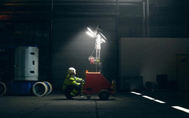 En person i varselkläder och hjälm justerar en röd mobil belysningsmast som lyser upp en mörk lagerlokal.