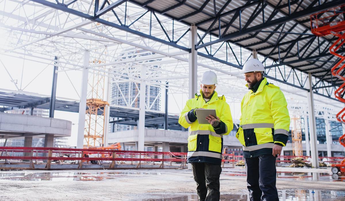 Två personer i gula varseljackor och vita hjälmar använder SitePlanner på en surfplatta inuti en stor stålkonstruktion under uppbyggnad.